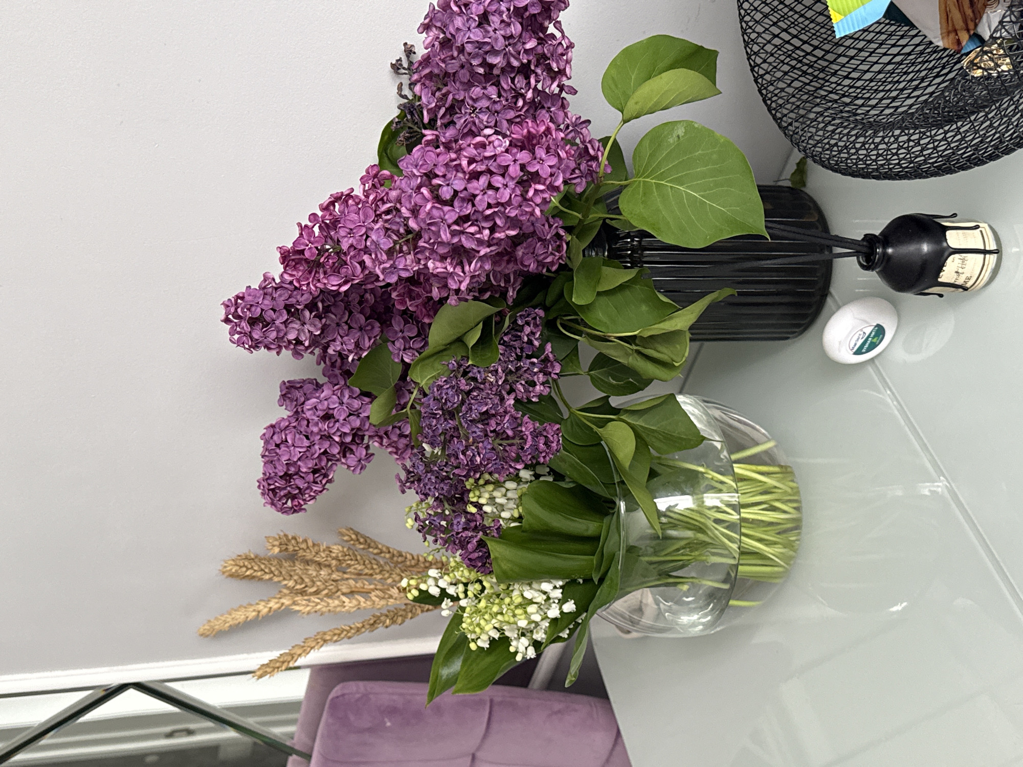 The image depicts an indoor still life arrangement, viewed from above, with the composition oriented vertically as if rotated clockwise.
**Subjects:** The primary subjects are a vibrant bouquet of purple lilac flowers, a cluster of delicate white lily of the valley, and several golden dried wheat stalks. The purple lilacs are partially held in a tall, black, ribbed vase. The white lily of the valley and wheat are displayed in a clear, spherical glass vase, revealing their green stems in water.
**Objects:** The floral arrangement rests on a glossy, light-colored table surface. In the upper right corner, a black woven wire basket contains indistinct items. Next to the black vase, there is a black reed diffuser bottle with a white label and several diffuser reeds. A small, white, oval-shaped object, possibly a soap bar or air freshener, is placed beside the diffuser. The background features a plain light grey wall, and a blurred section of a purple upholstered chair is visible in the lower left.
**Activity/Event:** No human activity is taking place; the image captures a static decorative composition.
**Notable details:** The setting is an indoor space, likely a living or dining area, characterized by clean lines and soft, even lighting. The glossy table surface reflects parts of the arrangement.
**Visible text:** Text is present on the white label of the black diffuser bottle and on a small green and blue label adhered to the white oval object. However, the text on both is too small and indistinct to be legibly identified.