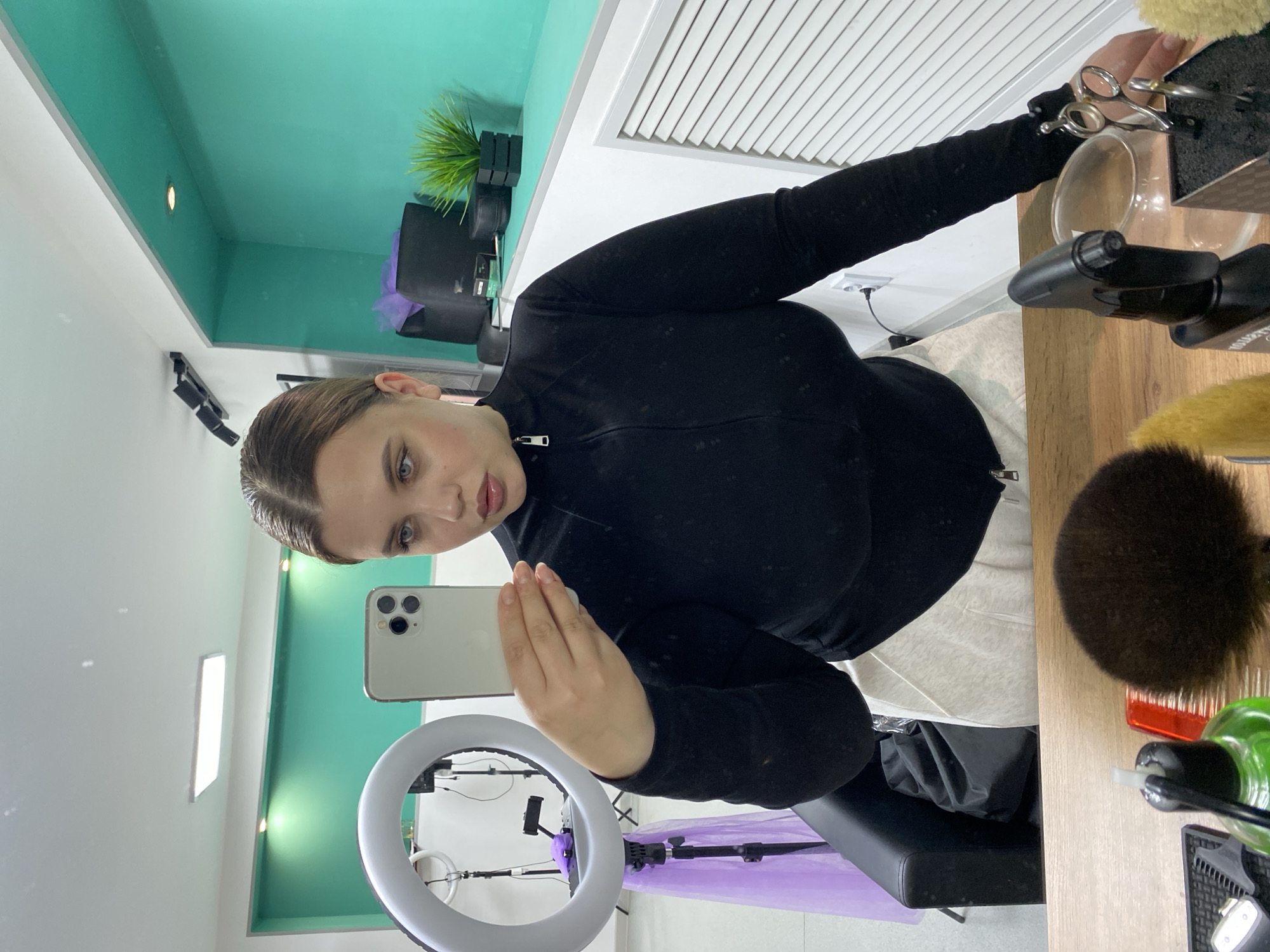 The image depicts a woman holding a smartphone, likely taking a selfie or recording a video, within an interior setting. She is positioned in front of a ring light and is dressed in a black, long-sleeved zippered top. The background includes a wall painted with a light green color and white ceiling panels. There is a plant visible in the top left. To the right, a table surface displays various items, including grooming tools like brushes and scissors. A black chair is visible beneath the woman. The location is Chernushka, Russia.