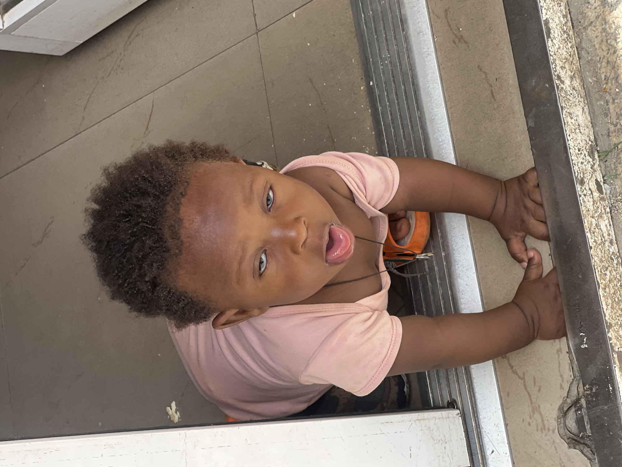 A young child with dark skin, curly hair, and light-colored eyes, wearing a pink shirt, is looking up at the camera with their tongue slightly out. The child is positioned on a beige tiled floor next to a metal frame, possibly a doorway or window frame. The child's hands are holding onto the metal frame. The location is Gura Top, Nigeria, based on the prompt. The background includes glimpses of what appears to be interior and exterior environments.