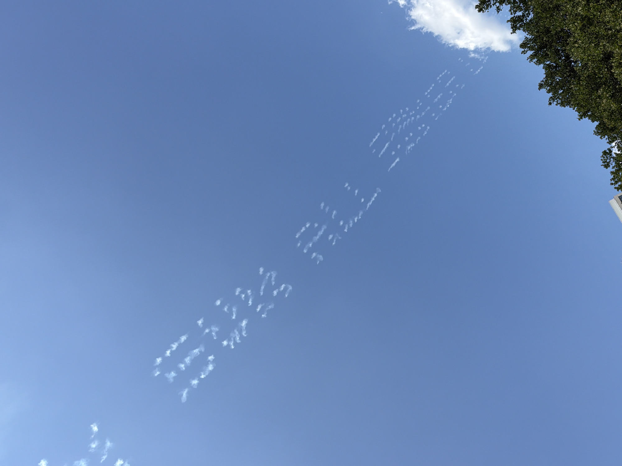 The image is a low-angle shot of a clear blue sky with some fragmented white clouds. In the upper left and diagonally across the sky, there are lines of small, dispersed clouds that appear to be in the shape of letters. The letters spell out "WE THE PEOPLE". The bottom of the image shows the top of a building and lush green trees. The scene is outdoors in a city environment.