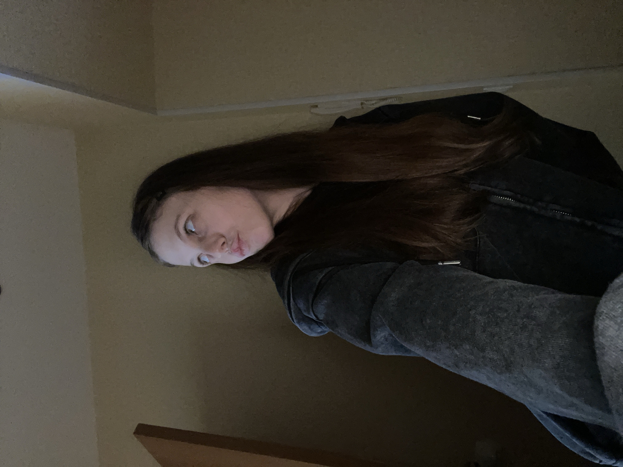 A young woman with long, dark brown hair is positioned in the center of the frame, looking up and to her left. She is wearing a dark gray, zip-up hoodie. Her face is angled, with her lips slightly pursed and her eyes directed upwards. The background consists of a dimly lit interior space, with walls that appear to be painted a muted yellow. To the left of the woman, a portion of a wooden door or cabinet is visible. The lighting is low, creating shadows and giving the scene a somewhat moody atmosphere. There are no discernible city landmarks or features in the image that would suggest a specific location within Ufa, Russia.