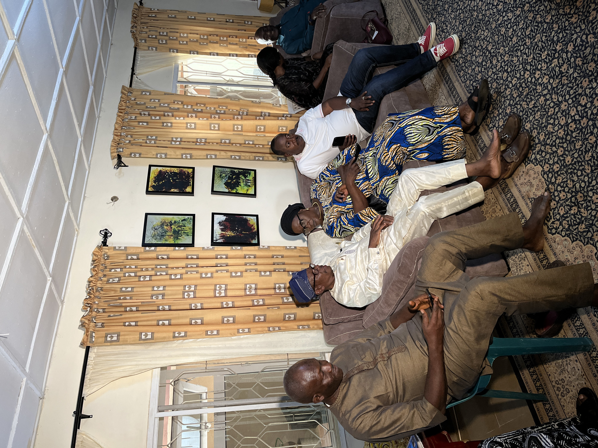 In a dimly lit indoor setting in Bukuru, Nigeria, a group of seven individuals is seated and standing. The room features patterned curtains, framed artwork on the walls, and a patterned carpet. The lighting suggests an evening or overcast day.
On the left, a man in a brown tunic and pants sits on a teal plastic chair, holding a dark mobile phone. Beside him, on a brown couch, a man wearing a white traditional outfit and a blue and gold patterned cap is seated, looking to the right. To his right, a woman in a vibrantly patterned blue, yellow, and orange dress is seated, her head bowed slightly. Next to her, a man in a white polo shirt and blue jeans is seated, holding a dark mobile phone. He is wearing a watch on his left wrist. To his right, a woman in a black patterned top is seated, her head down. Further to the right, a man in a teal-colored shirt is seated and smiling, looking towards the left. Partially visible on the far right, another individual in dark clothing is seated.
The individuals appear to be engaged in conversation or observing something off-camera. The overall atmosphere is relaxed and informal. The decor and attire suggest a comfortable, domestic environment.