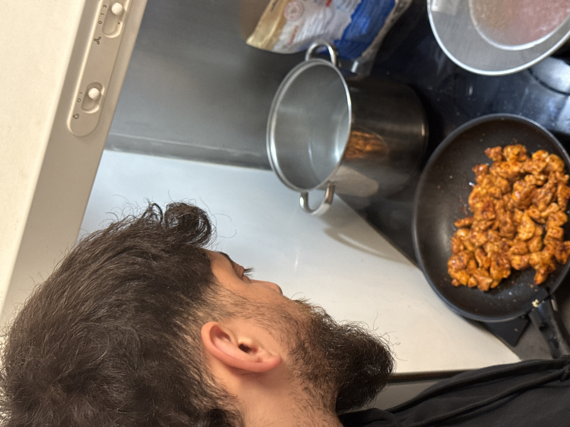 A close-up, high-angle shot depicts a man with dark hair and a beard looking upwards towards a kitchen counter and stovetop. On the stovetop, there's a pot and a frying pan filled with cooked, orange-colored food. A white range hood is visible above the counter. The city of Strasbourg, France is not discernable from the visual information provided.