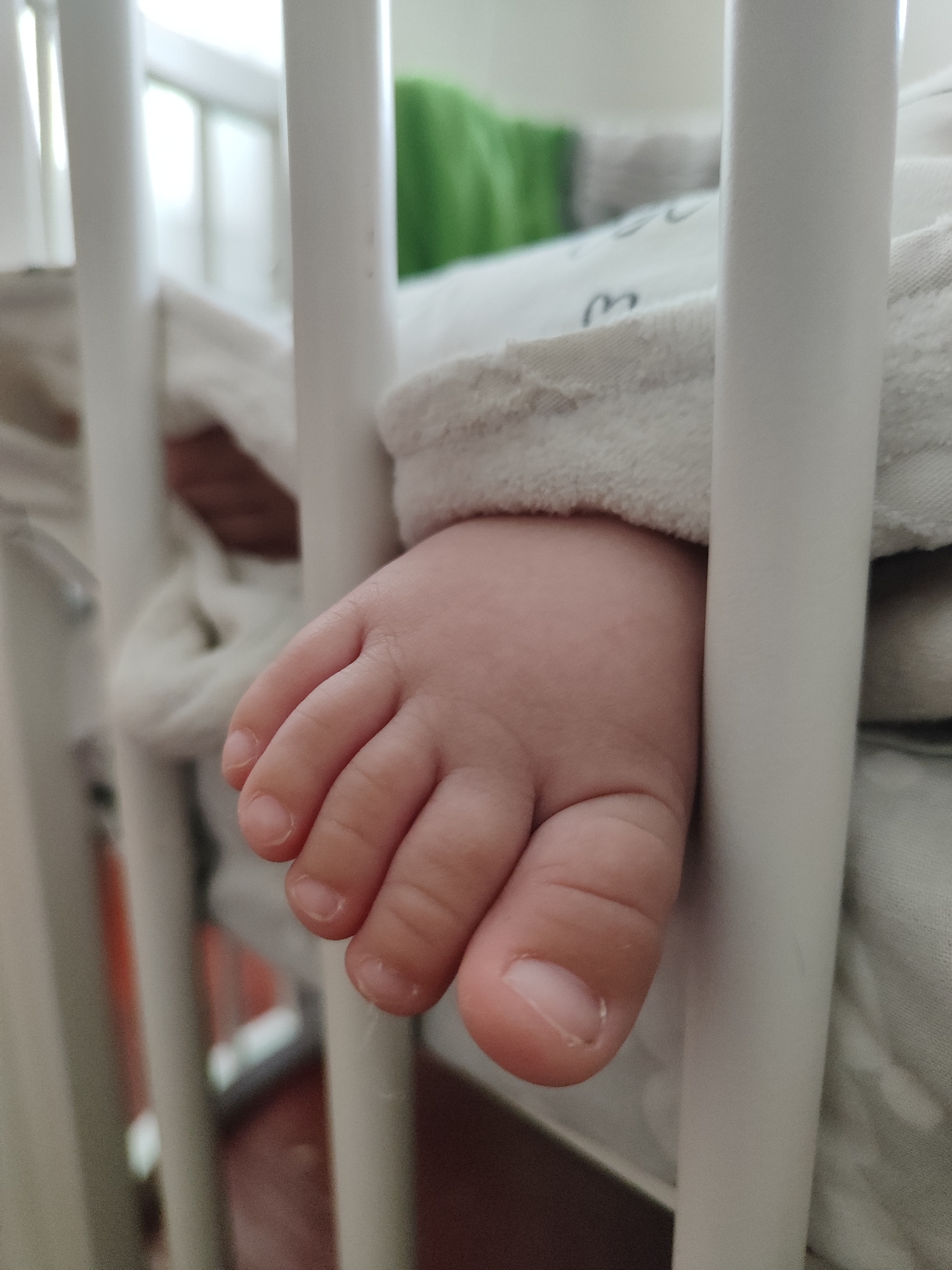 The image is a close-up shot of a baby's foot peeking out from under a blanket in a crib. The baby's skin is smooth and pink, and its toes are clearly visible, with tiny fingernails. The foot is resting against the white bars of the crib, and a soft, white blanket is partially covering the baby. In the background, a blurred green object can be seen, possibly a toy or a piece of clothing. The scene is brightly lit, suggesting it could be daytime. The overall impression is one of tenderness and innocence. There is no visible text in the image.