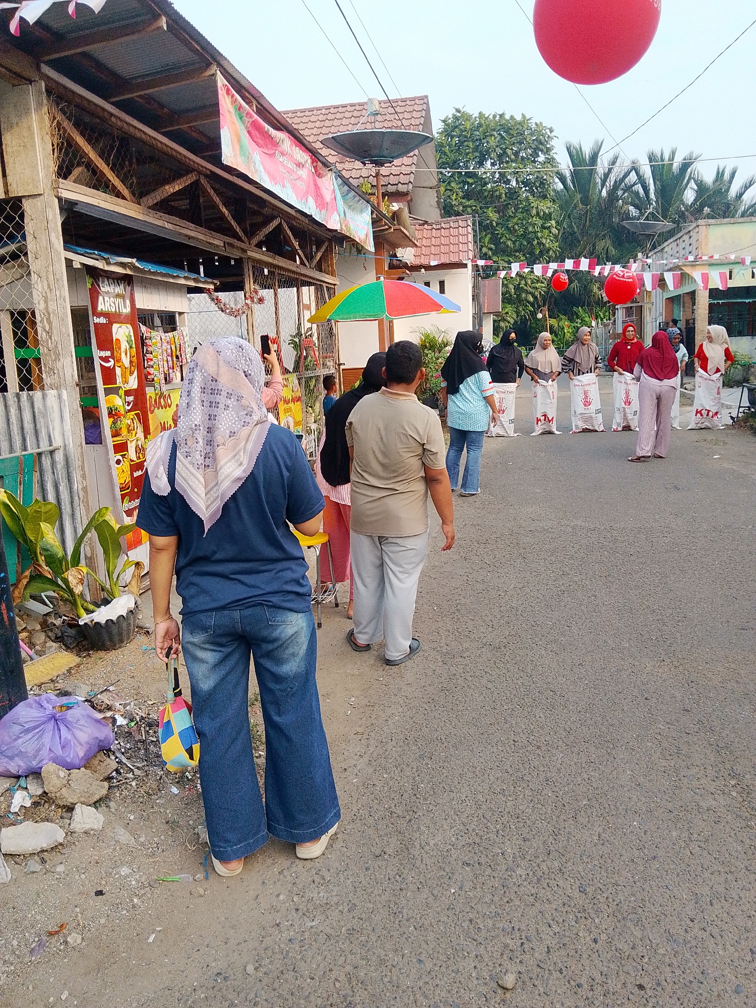 A group of women, some wearing hijabs, stand in a street in Barabai, Indonesia, participating in a sack race. They are lined up behind large white sacks marked with text. Red balloons are visible in the sky above, along with festive red and white Indonesian flags. The street is paved and lined with buildings and trees. In the foreground, a woman in jeans and a blue shirt is visible from behind, holding a colorful bag. Trash is scattered on the side of the road.