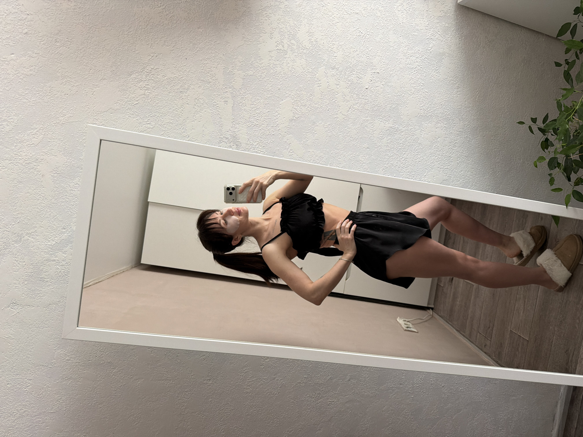 A woman is captured in a mirror selfie, posing in what appears to be an indoor setting, likely in Perm, Russia. She is seen from the waist up in the mirror's reflection, with her lower legs and feet also visible.
The woman has dark hair styled in a low ponytail with bangs. She is wearing two white under-eye patches, suggesting a skincare routine. Her outfit consists of a black ruffled crop top with thin straps and black shorts or a mini skirt. She is also wearing brown, fuzzy slippers. Her left arm is raised, holding a silver smartphone with multiple cameras, indicating she is taking the mirror selfie. Her right hand is placed on her hip, slightly adjusting her clothing. A tattoo is visible on her side/ribcage area.
The large, rectangular mirror has a white frame and is propped against or mounted on a textured, off-white wall. The reflection shows parts of a room, including white cabinets or furniture, a light-colored floor, and a green potted plant on the right side. An electrical outlet with white cords is visible on the wall reflected in the lower right corner. The lighting in the room is bright and even. There is no visible text in the image.