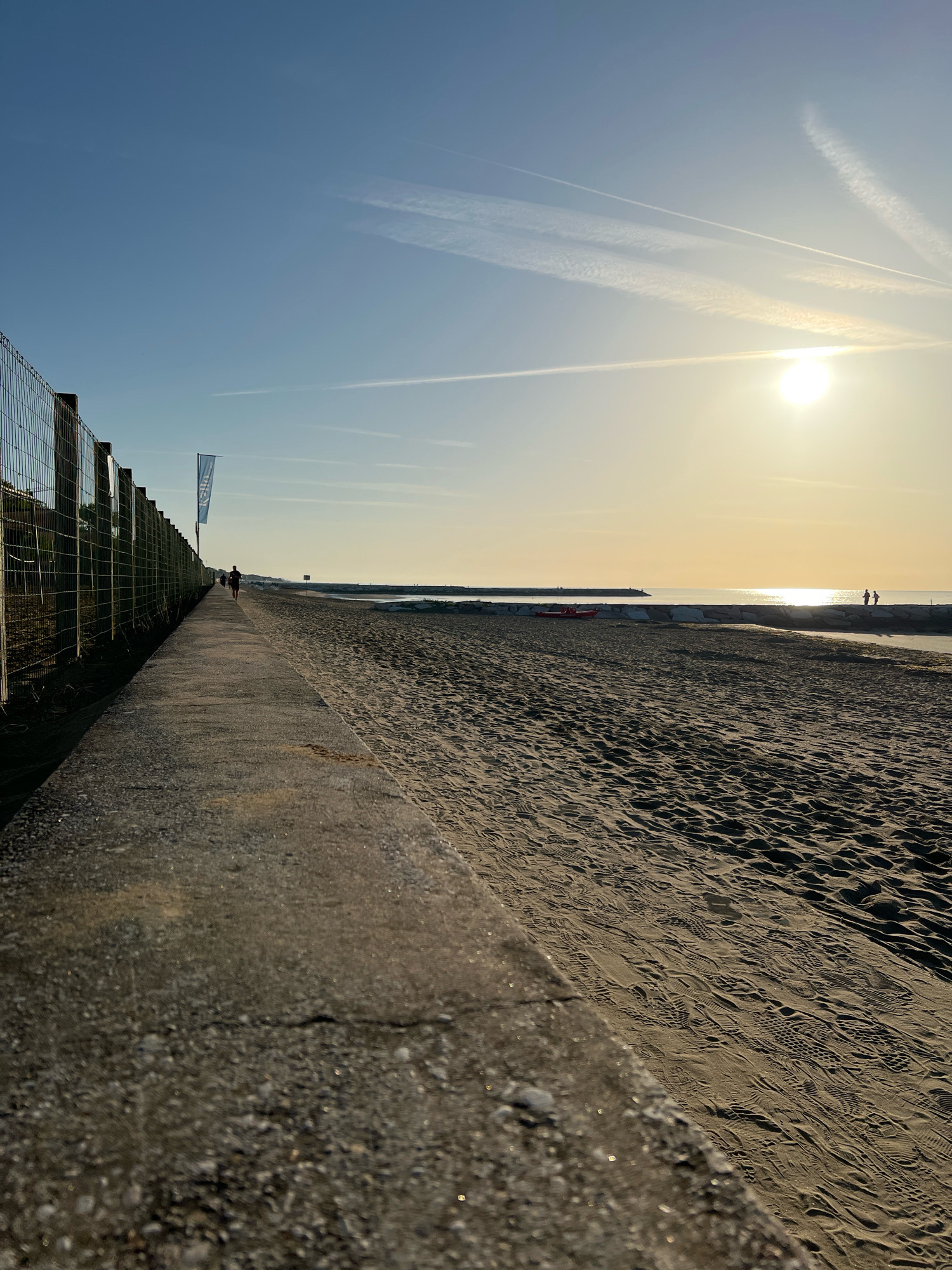 A low-angle, wide shot captures a beach scene at sunrise in Caorle, Italy. A concrete walkway runs along the left side of the image, bordering a sandy beach. A chain-link fence is visible along the edge of the walkway. In the distance, a lone figure walks along the walkway, and further down the beach, two figures stand near the water. The sun is low in the sky, casting a warm glow over the scene and creating reflections on the water. There are contrails in the sky, suggesting airplane activity. The sand is marked with numerous footprints, indicating recent activity. A red rescue boat is visible on the beach near a stone breakwater.