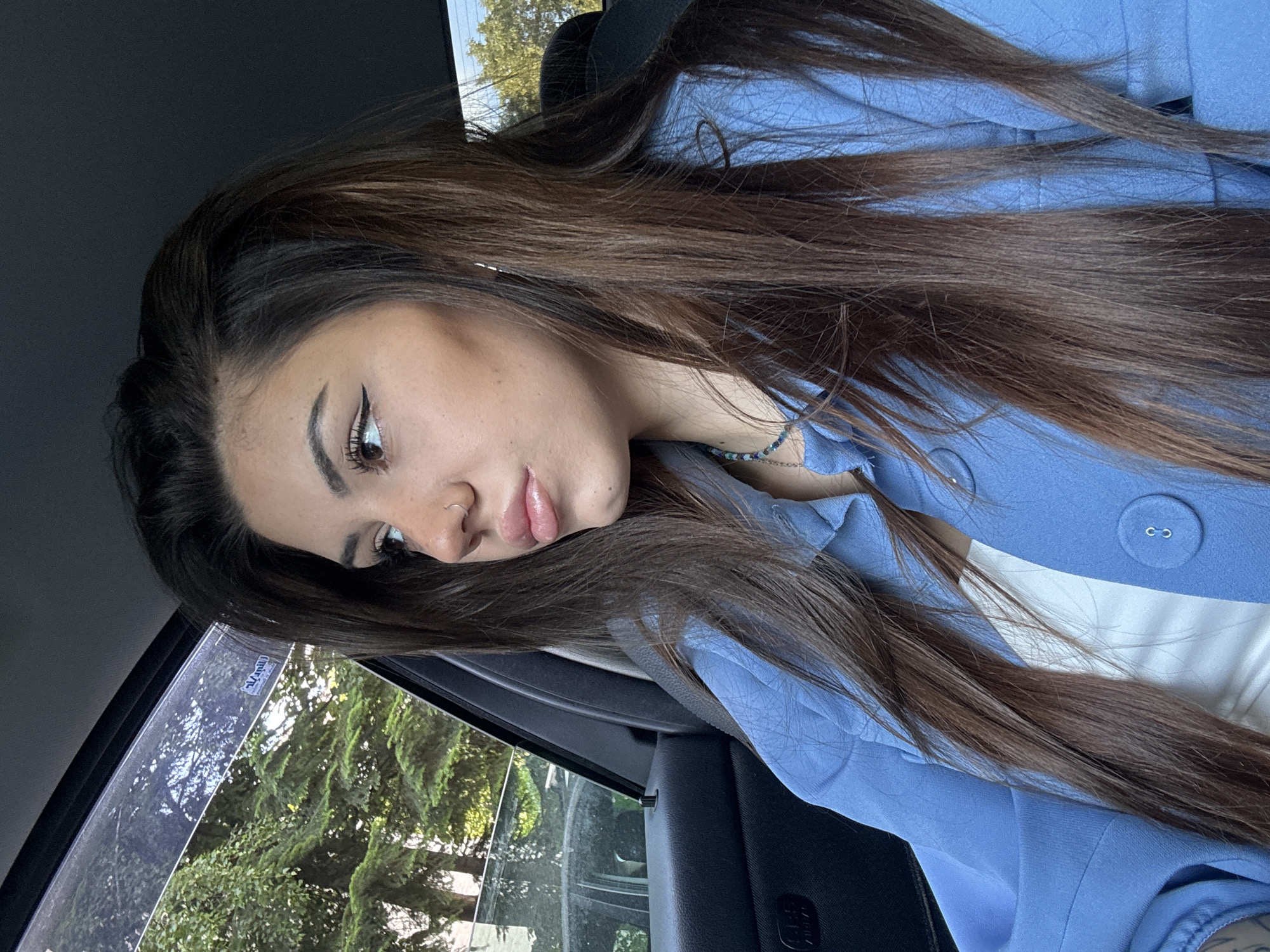 The image, rotated 90 degrees clockwise, features a young woman with long, flowing dark brown hair. She is situated inside a car, with her body angled and her head slightly tilted downwards, exhibiting a pensive or calm expression. Her visible makeup includes prominent winged eyeliner and defined eyebrows, complementing her light brown or hazel eyes. A small silver hoop nose piercing is visible on her left nostril, and her lips are a soft pink.
She is wearing a light blue blazer or jacket over a white inner top. A round, fabric-covered button is visible on the blazer. A delicate beaded bracelet with blue and green beads adorns her right wrist.
The setting is clearly inside a vehicle, with parts of a dark car seat or interior panel visible. Through the car window on the left, lush green trees and foliage are seen under natural daylight, suggesting an outdoor environment, possibly a street or park in a location like Sofia, Bulgaria. The lighting indicates it is daytime with clear weather. A small, rectangular sticker on the car window bears the text "Vannill." The overall activity appears to be a posed photograph or a relaxed moment while traveling in a car.