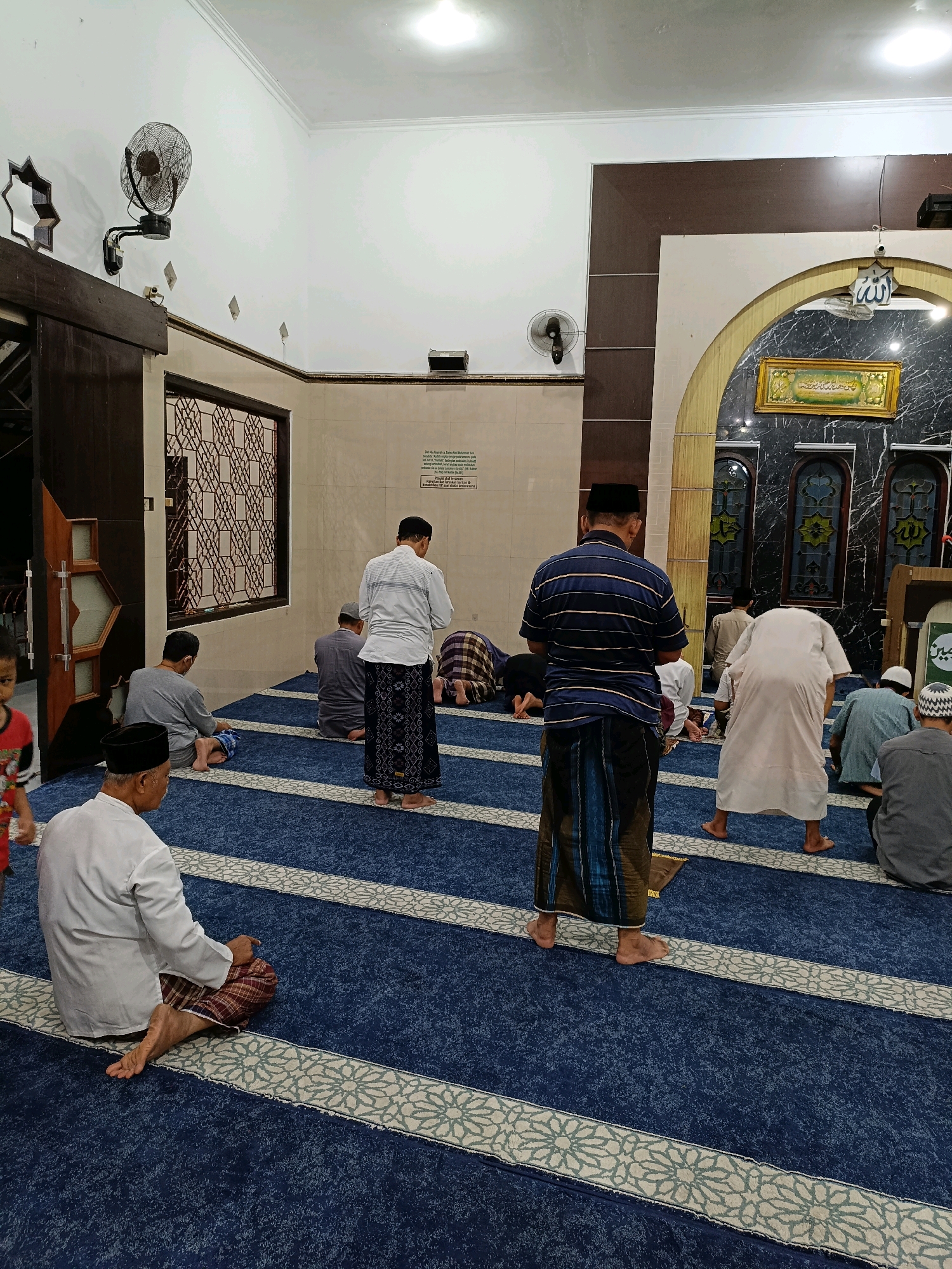 The image depicts the interior of a mosque in Maguwoharjo, Indonesia, with several individuals engaged in prayer. The flooring is covered with a blue carpet featuring a repeating white and green pattern. In the foreground, a man in a white tunic and a checkered sarong is seated on the carpet. Behind him, a man in a blue and white striped shirt and a sarong stands facing away from the camera. Several other men are visible in various prayer postures, kneeling or bowing, some wearing traditional Islamic attire. A young child in a red and black shirt stands to the far left. The mosque's architecture includes an arched doorway with ornate detailing, a framed artwork with Arabic calligraphy, and patterned windows. A wall-mounted fan is visible on the left side of the image, and light fixtures are mounted on the ceiling. The overall scene suggests a communal religious gathering.