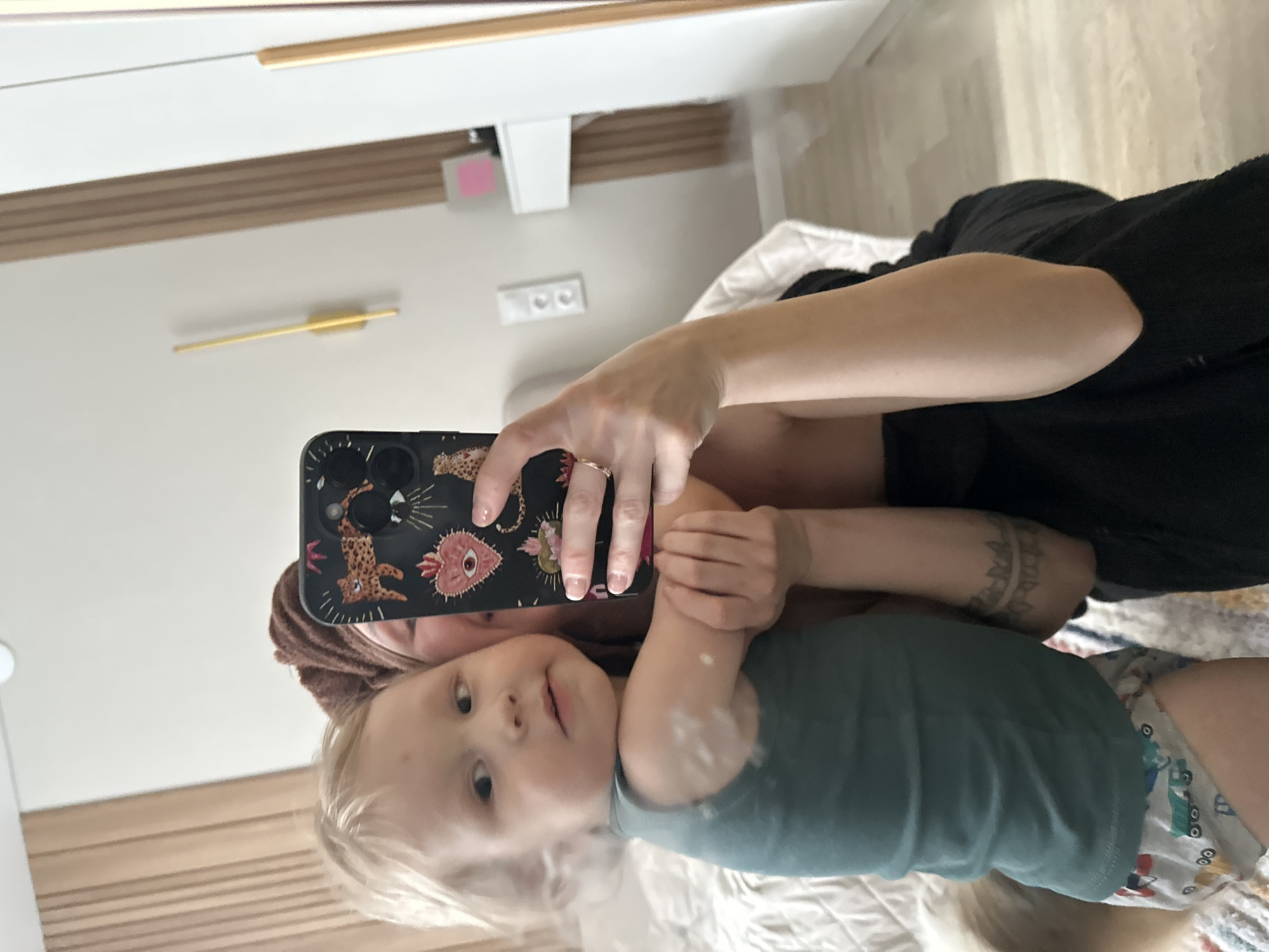 A woman in a black robe holds a child with blond hair, both reflected in a mirror. The woman is using a black phone with a decorated case to take a selfie. The child wears a green shirt and patterned diaper. The background includes a light-colored bed, a white wall with a clock, and electrical outlets. The location is likely indoors, possibly a residence in Votkinsk, Russia.