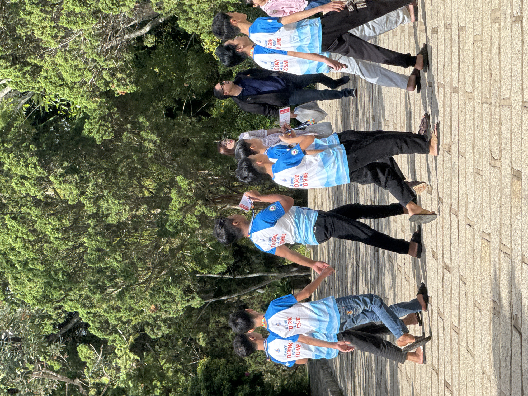 This image captures a group of individuals, predominantly young males, walking along a paved stone path lined with lush green cypress trees. The setting appears to be a park or an outdoor venue in Da Lat, Vietnam, indicated by the dense coniferous vegetation characteristic of the region's highlands.
The individuals are mostly wearing matching blue and white t-shirts with a wave-like design on the bottom and a logo on the left chest. Some are holding printed materials. Their attire and the synchronized nature of some of the t-shirts suggest they might be part of a school group, a team, or a similar organized gathering.
The group is walking from left to right in the frame. On the far left, two young males in similar blue and white t-shirts are walking closely together. To their right, another young male in the same t-shirt is walking, also holding a pamphlet. Further to the right, a group of three young males in the same t-shirts are walking in a line, one of them holding a pamphlet and the other two engaged in conversation. In the background, an adult male in a black jacket and dark pants is walking, holding a white plastic bag. To his left, an adult female in a light-colored patterned dress is standing and looking towards the group.
The environment is dominated by the vibrant green of the cypress trees, which form a dense canopy overhead, creating a dappled light effect on the path. The stone path is made of rectangular slabs and shows signs of wear, consistent with a well-trafficked area. The lighting suggests it is daytime, with the sun casting shadows on the ground. The overall atmosphere is relaxed and casual, indicative of a leisurely walk or a group outing.