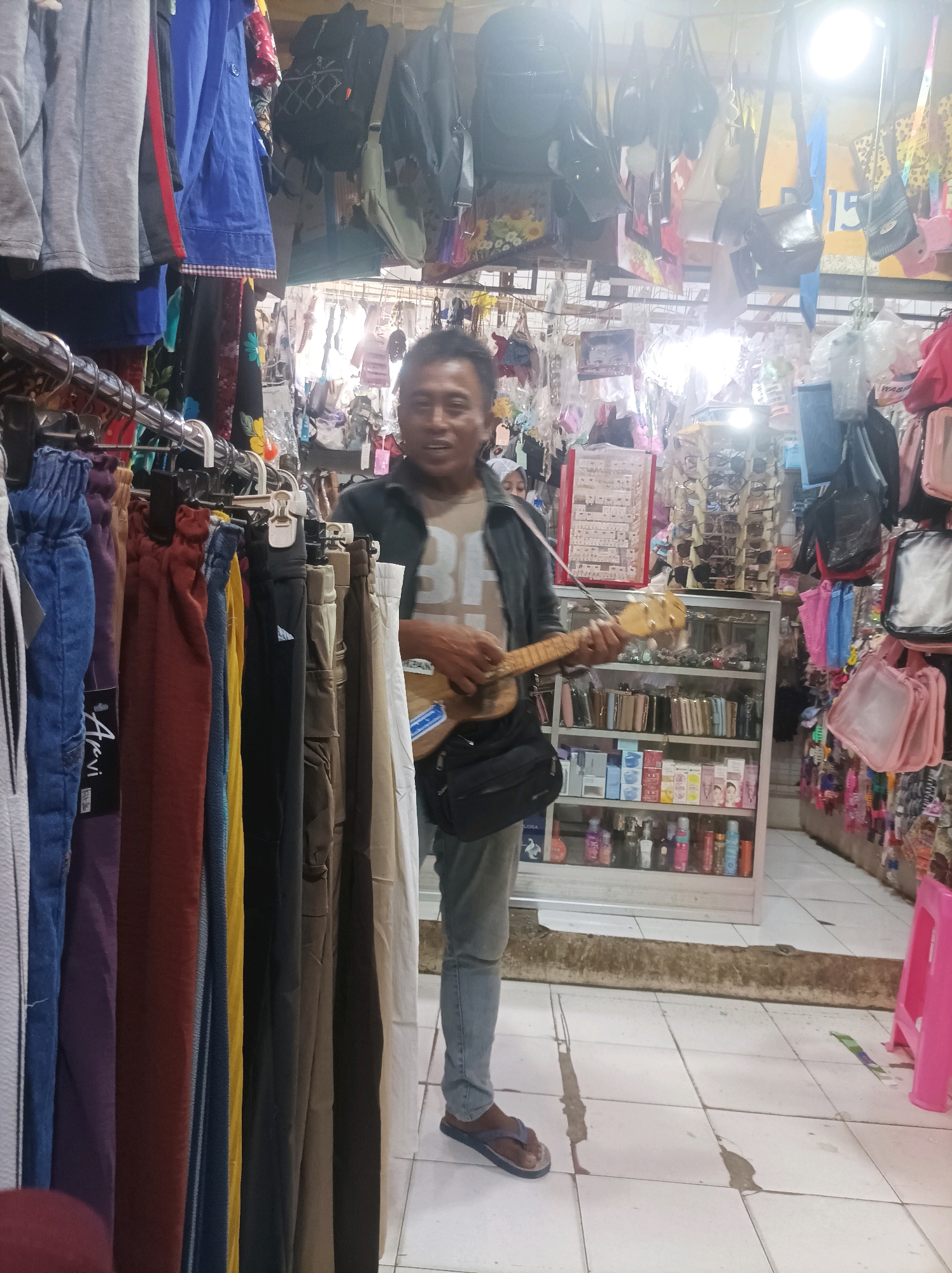 The image captures a man standing in a market stall, holding a small guitar and appearing to play it. He is wearing a light brown t-shirt with a graphic design and blue jeans. A black bag is slung across his chest. The stall is filled with various items for sale, including racks of clothing such as pants and jackets, as well as shelves displaying merchandise like bags, accessories, and toiletries. In the background, more items are hung from the ceiling, including bags and clothing. The floor is tiled, and a pink plastic stool is visible to the right. The scene suggests a lively, perhaps informal, marketplace setting within Sumuragung, Indonesia.