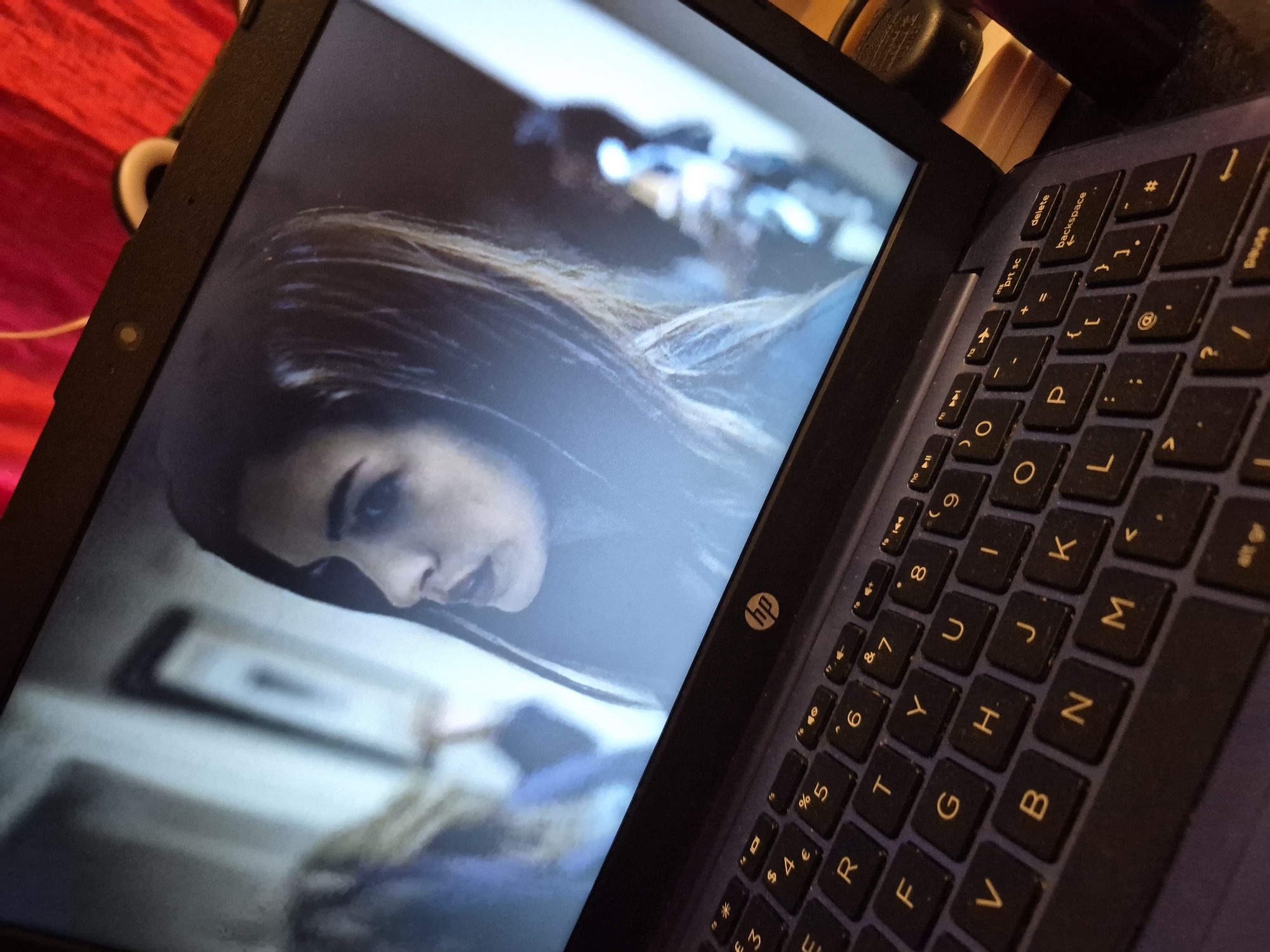 The image is a close-up, high-angle shot of a laptop computer screen displaying a scene with a woman's face, partially in focus. The laptop is open, and a portion of its keyboard is visible in the foreground. The keyboard is black with yellow lettering. The screen shows a woman with dark hair, her face turned to the left, with her mouth slightly open. The background on the screen is blurred and appears to be an indoor setting with light-colored walls and some indistinct figures. The overall lighting is dim, with a reddish glow from a light source outside the frame, possibly fabric. The laptop itself is dark blue. There is no discernible location in Sandwell, United Kingdom.