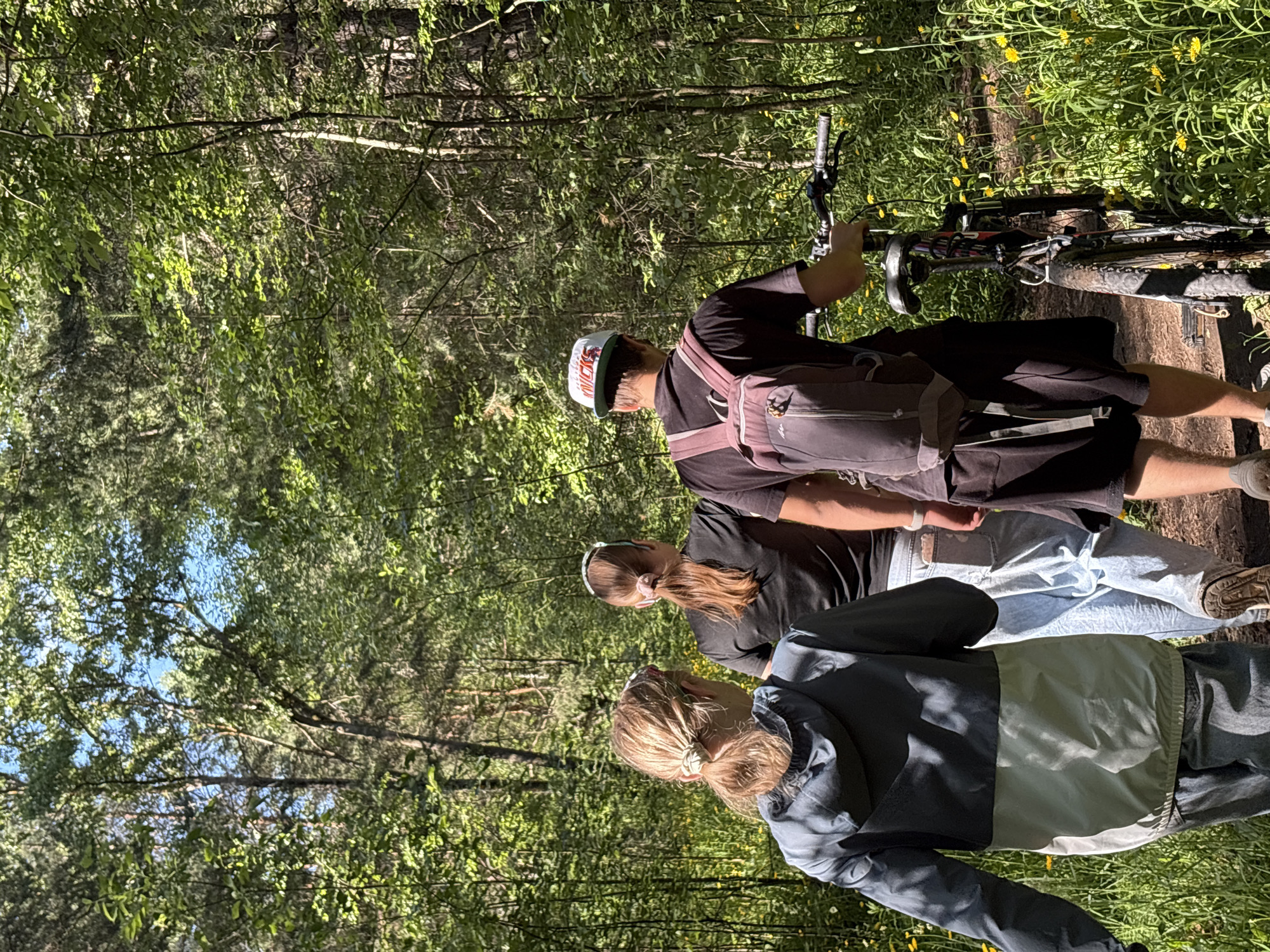 Three individuals, two women and one man, are walking along a dirt path in a sun-dappled forest. The woman in the foreground on the left wears a two-toned jacket, grey and light green, and her hair is pulled back in a ponytail. The middle woman, wearing a black t-shirt and jeans, has her brown hair in a ponytail and wears a patterned headband. The man on the right is wearing a black t-shirt, black shorts, a white baseball cap, and a maroon backpack. He is pushing a bicycle with black tires and a red frame. The path is lined with green grass and small yellow flowers. The trees surrounding the path are tall and green, with sunlight filtering through the leaves, creating dappled light and shadows on the ground. The scene appears to be from a recreational activity in a natural setting, likely a park or trail within or near Chaikovsky, Russia, given the context.