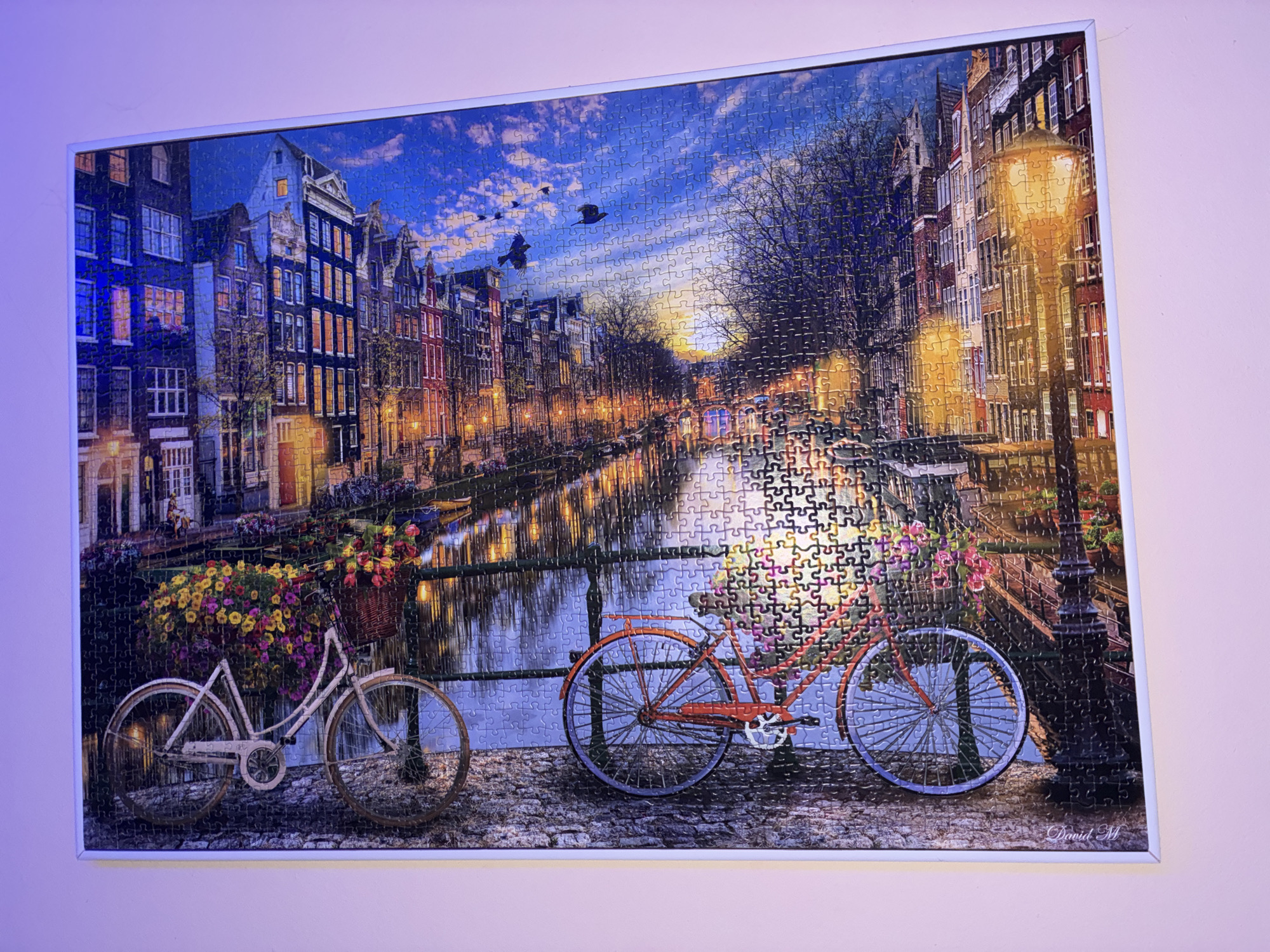 This is a framed jigsaw puzzle depicting a picturesque canal scene in Amsterdam, Netherlands. The puzzle is comprised of numerous interlocking pieces that together form a detailed image. The scene captures a canal lined with traditional Dutch canal houses, their windows illuminated with warm, inviting light. Two bicycles are prominently featured in the foreground, parked on a cobblestone path. The bicycle on the left is white and has a basket overflowing with colorful flowers, while the bicycle on the right is red and has a similar floral arrangement. A lamppost with a glowing light stands near the red bicycle, casting an orange hue on the surrounding area. The water of the canal reflects the lights of the houses and the sky, which is a deep blue with scattered clouds and birds in flight. The overall impression is one of a tranquil and charming evening in a historic European city.