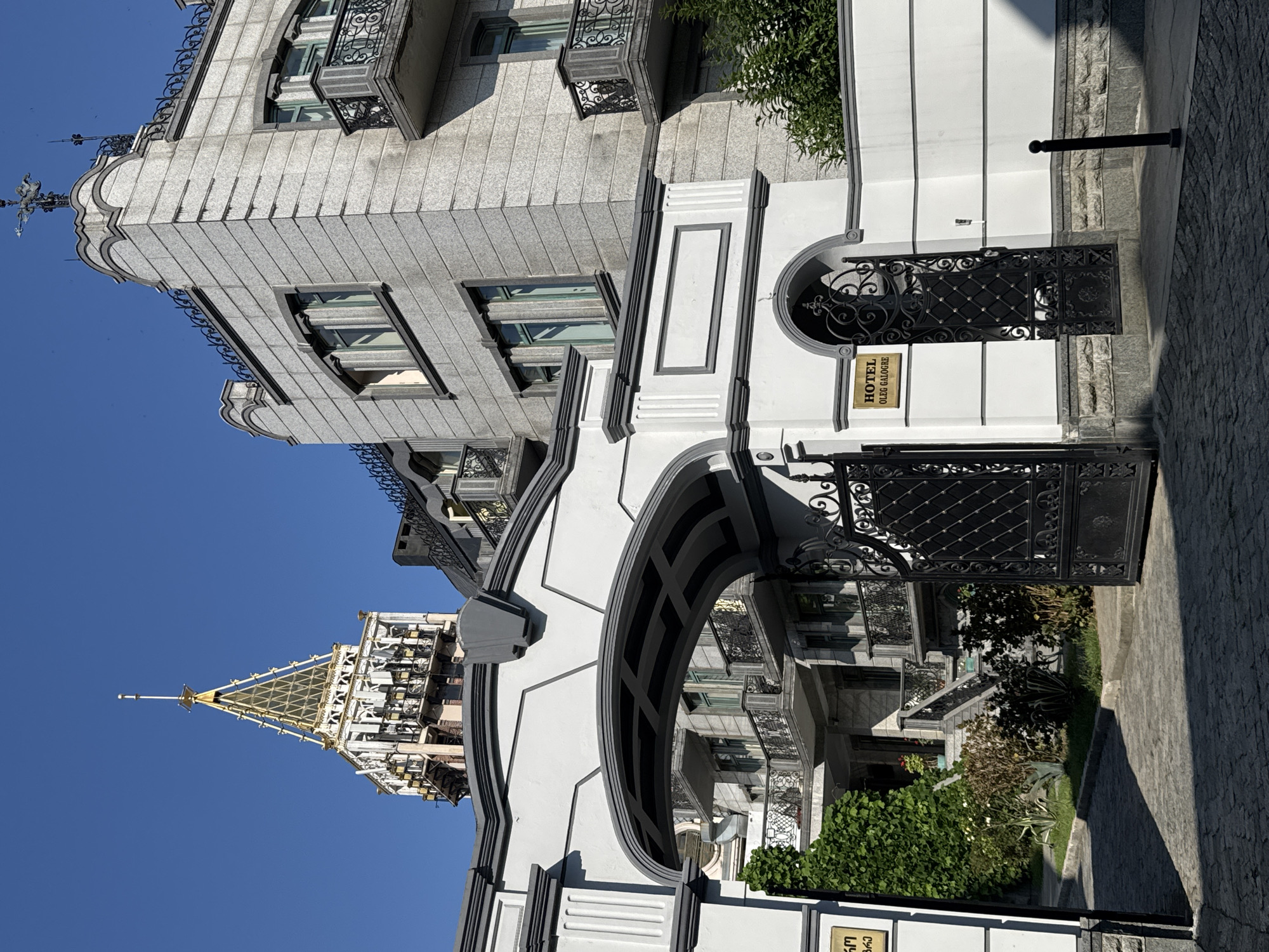 In Batumi, Georgia, a grand, ornate white archway with dark grey detailing leads into a courtyard. Through the arch, a cobblestone path is visible, lined with lush green bushes and ornamental plants. Beyond the arch, a building with grey stone facade, multiple balconies with black wrought-iron railings, and tall windows with green-tinted glass stands bathed in sunlight. In the distance, a distinctive tower with a golden, pyramidal roof and white architectural elements pierces the clear blue sky. A dark metal gate, adorned with intricate scrollwork, stands open on the right side of the archway, next to a small plaque that reads "HOTEL OLEG GALOGRE". The scene appears to be a sunny day in a city known for its coastal architecture and unique landmarks.