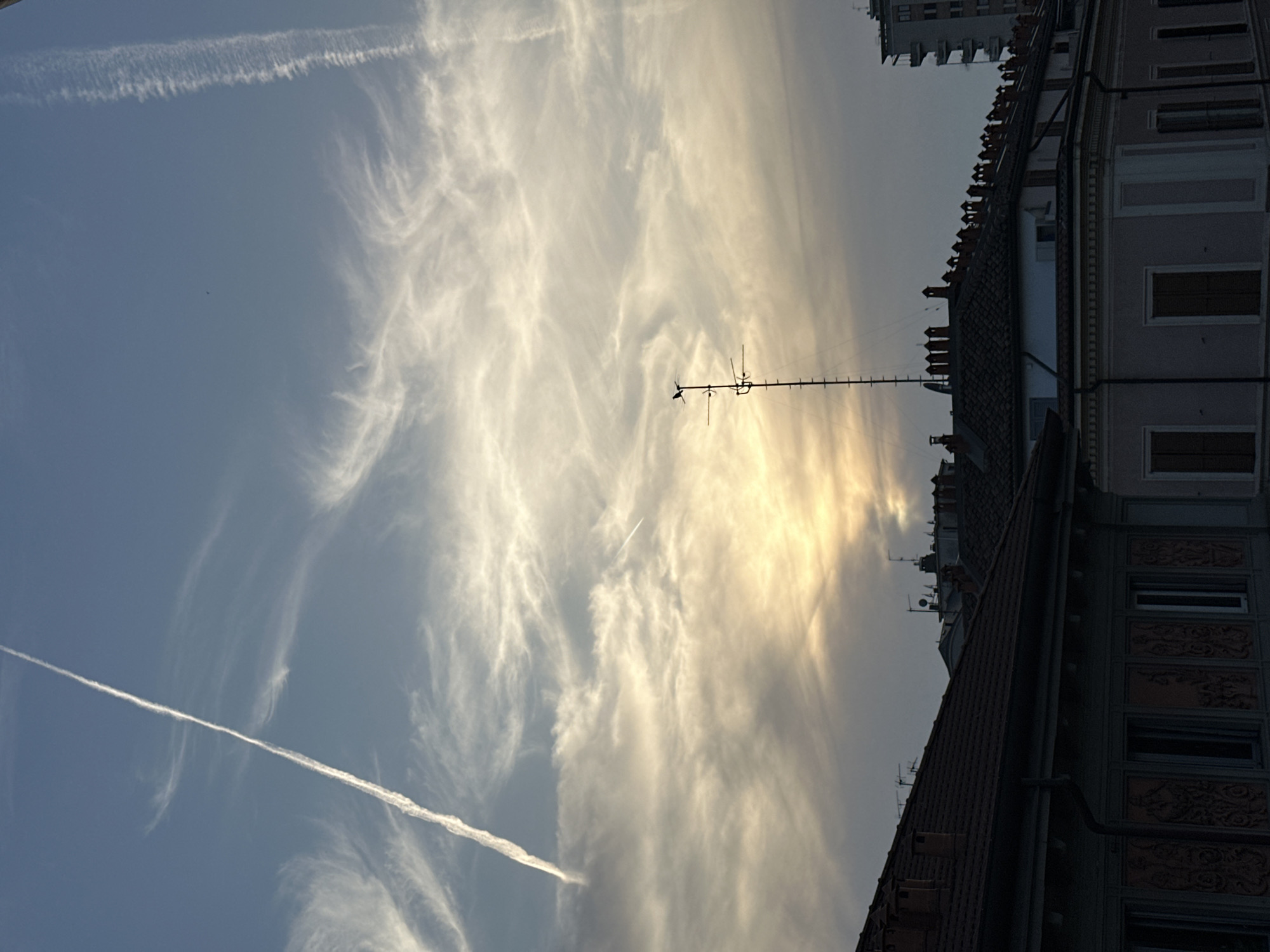 The image shows a view of the sky over Milan, Italy. In the foreground is a building with a red-tiled roof and multiple chimneys. A television antenna is visible on the roof. Behind this building, there is another building with windows and decorative panels. The sky is filled with wispy clouds and contrails, with a golden glow suggesting the sun is low on the horizon. The light and clouds dominate the scene.