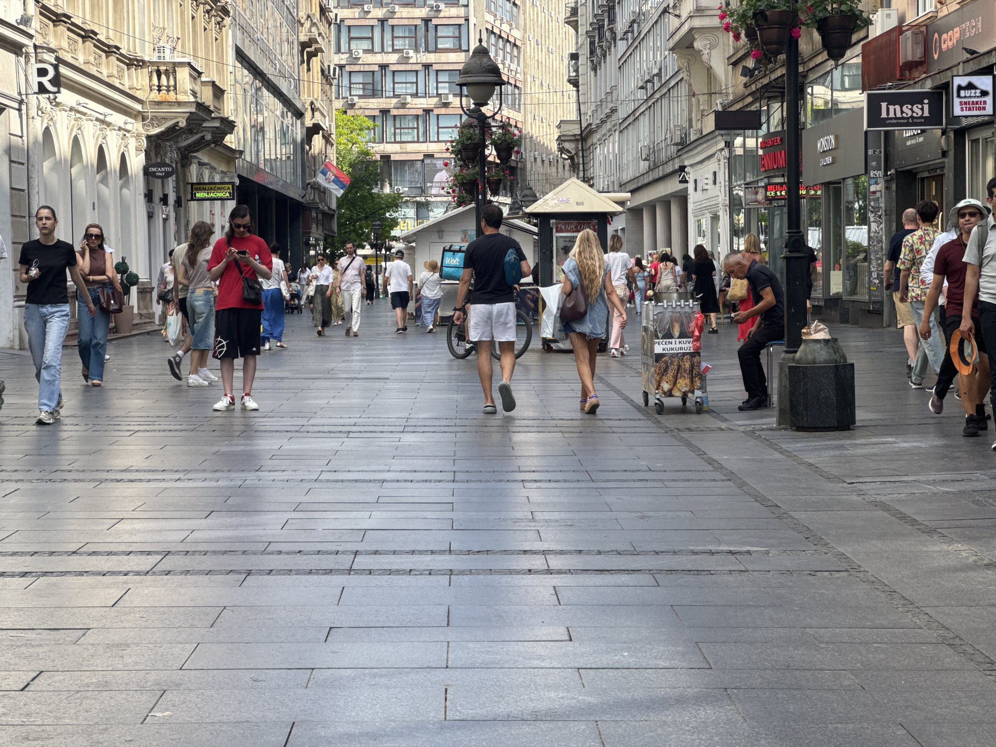 This media file captures a bustling pedestrian street in Belgrade, Serbia. The scene is filled with people walking in various directions, suggesting a busy day. Several individuals are the main subjects, including a woman in a black t-shirt and jeans, a man in a red t-shirt and shorts looking at his phone, and a woman with blonde hair in a denim dress.
The environment is an urban street lined with shops and buildings. Visible signs include "INCANTO ITALY," "MENJAČNICA" (exchange office), "POPCORN," "MONSOON CHILDREN," "Inssi underwear & more," and "BUZZ SNEAKER STATION." There's also a sign in Cyrillic that reads "PEČEN I KUVA KUKURUZ," likely indicating roasted and boiled corn for sale. A Serbian flag is prominently displayed on one of the buildings.
The time of day appears to be daytime, with clear shadows indicating sunlight. The weather seems pleasant, suitable for walking outdoors. The overall mood is one of everyday activity and commerce, with people engaged in shopping, commuting, or simply enjoying a stroll. The paved ground and architectural details of the buildings contribute to the urban setting. The presence of shopping bags and people in casual attire reinforces the idea of a commercial district.