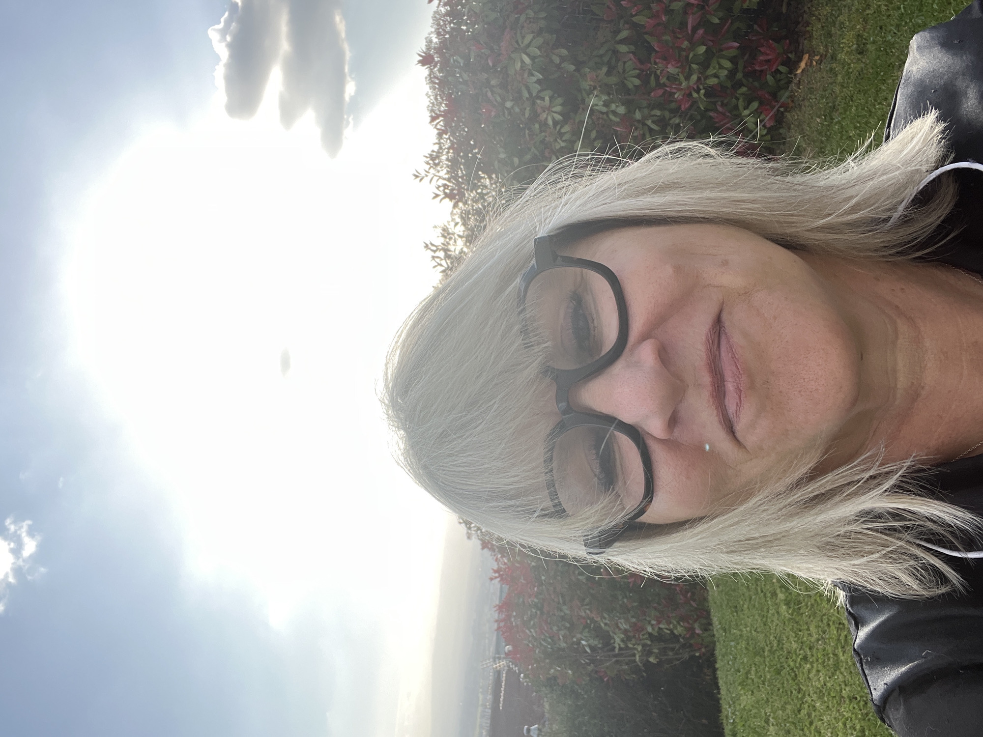 A selfie portrait shows a blonde-haired woman wearing black-rimmed glasses. She is positioned in front of a background of green grass and a tree with red leaves. In the distance, a cityscape is faintly visible under a bright sky with clouds. The city is Chambœuf, France.