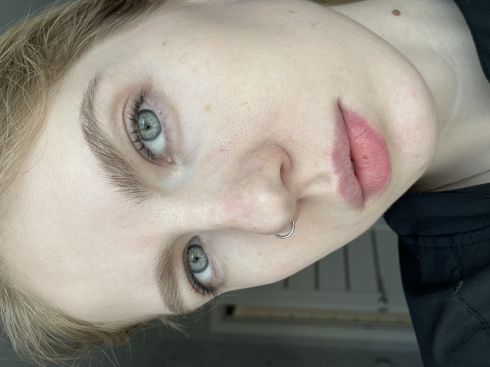 This image captures a close-up, head-and-shoulders portrait of a single female individual, set indoors in Izhevsk, Russia. The subject has fair skin, light blonde hair visible at the temples and forehead, and light blue-green eyes. Her eyebrows are naturally shaped and light brown. A silver-colored, circular septum piercing is present on her nose. Her lips are a natural pink tone and slightly parted. Fine skin texture, including visible pores, small freckles or beauty marks, and subtle localized redness around the nasal bridge and central forehead, is discernible. A prominent, raised, light brown mole is located on the right side of her neck, just below the jawline. She is wearing a dark, possibly black, collared garment, with a thin, silver-colored chain necklace partially visible. The individual is looking directly towards the camera. The background is out of focus, appearing as a dark gray indoor environment, with a faint white door or doorframe outline visible on the left side of the frame. The lighting is soft and frontal.