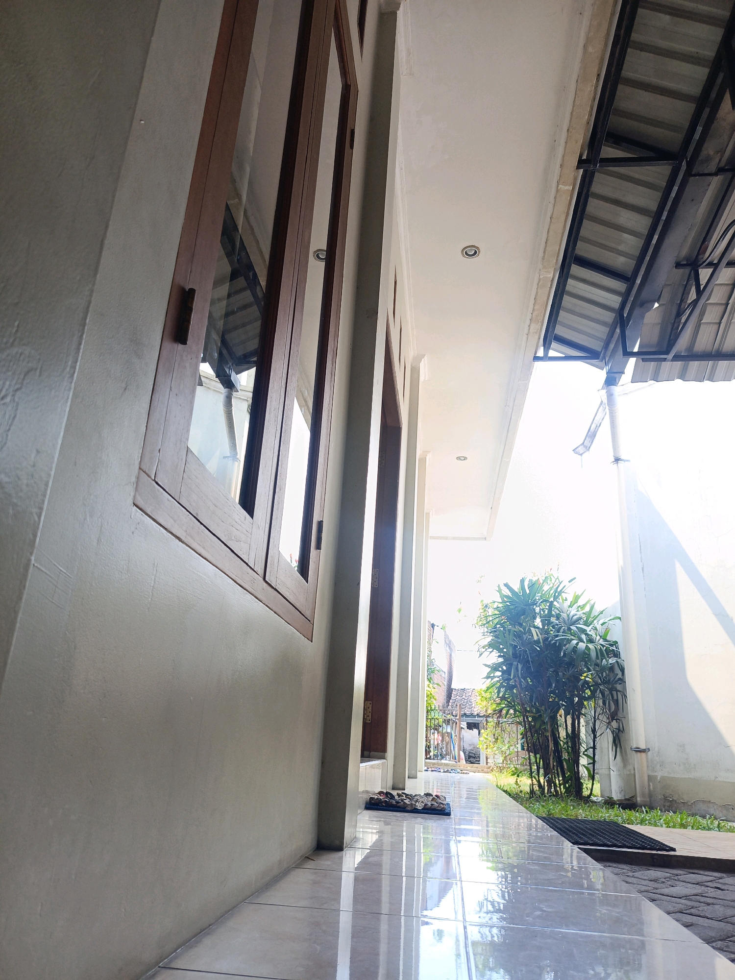 The image captures a narrow pathway alongside a building with a tiled floor that reflects the overhead sunlight. The building on the left has a light-colored wall and a wooden-framed window. To the right, a white wall borders the pathway, and an overhead structure with a corrugated metal roof is visible. In the background, a cluster of green leafy plants grows, and a glimpse of other buildings and a fence suggests a residential area. A drainpipe runs down the white wall. There are no people visible in the image. The scene appears to be an outdoor walkway in a warm, sunny climate, possibly in Magetan, Indonesia, given the architectural style and vegetation.
