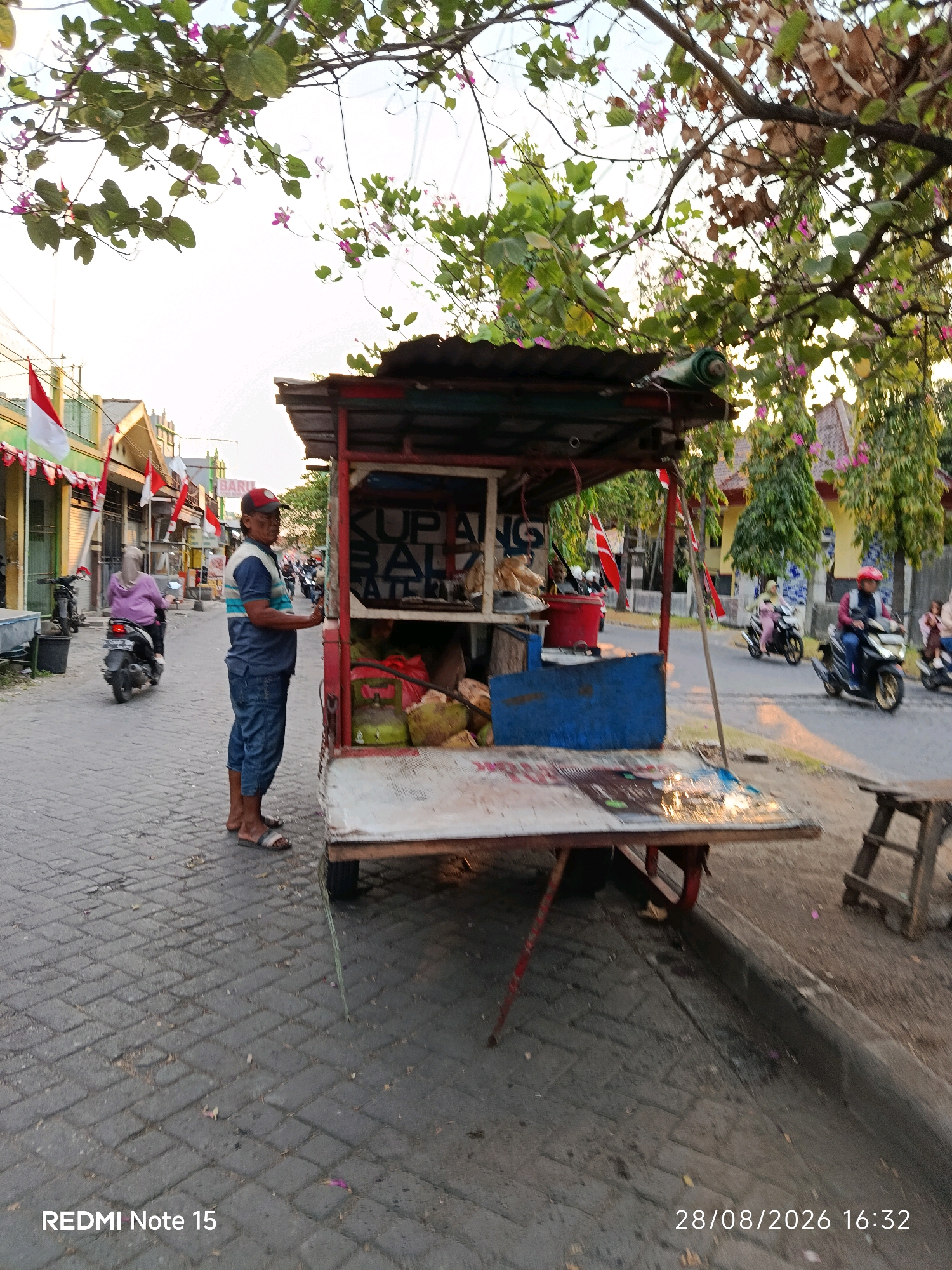 In the city of Keboansikep, Indonesia, a man stands beside a mobile food cart under the shade of a tree. The cart, made of wood and corrugated metal, displays coconuts and other items. The man, wearing a red cap, blue shirt, and denim shorts, appears to be a vendor. In the background, a woman rides a motorcycle, and other people and vehicles are visible on a paved street lined with buildings adorned with Indonesian flags. The scene captures a typical street view in a local Indonesian community.