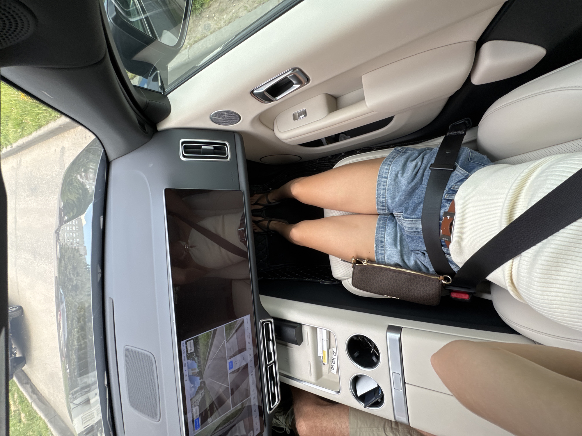 An overhead shot looks into the passenger side of a car, showing the interior and some of the exterior. The car is in an outdoor, urban location on a paved surface. In the foreground, a person's legs and torso are visible, wearing denim shorts and a white ribbed top. The person is wearing a seatbelt. A small brown designer purse is clipped to the seatbelt. The car's dashboard is in view, with a large infotainment screen displaying a camera feed of the exterior. The interior of the car is light beige and black. Outside the car, green grass and a curb are visible.
