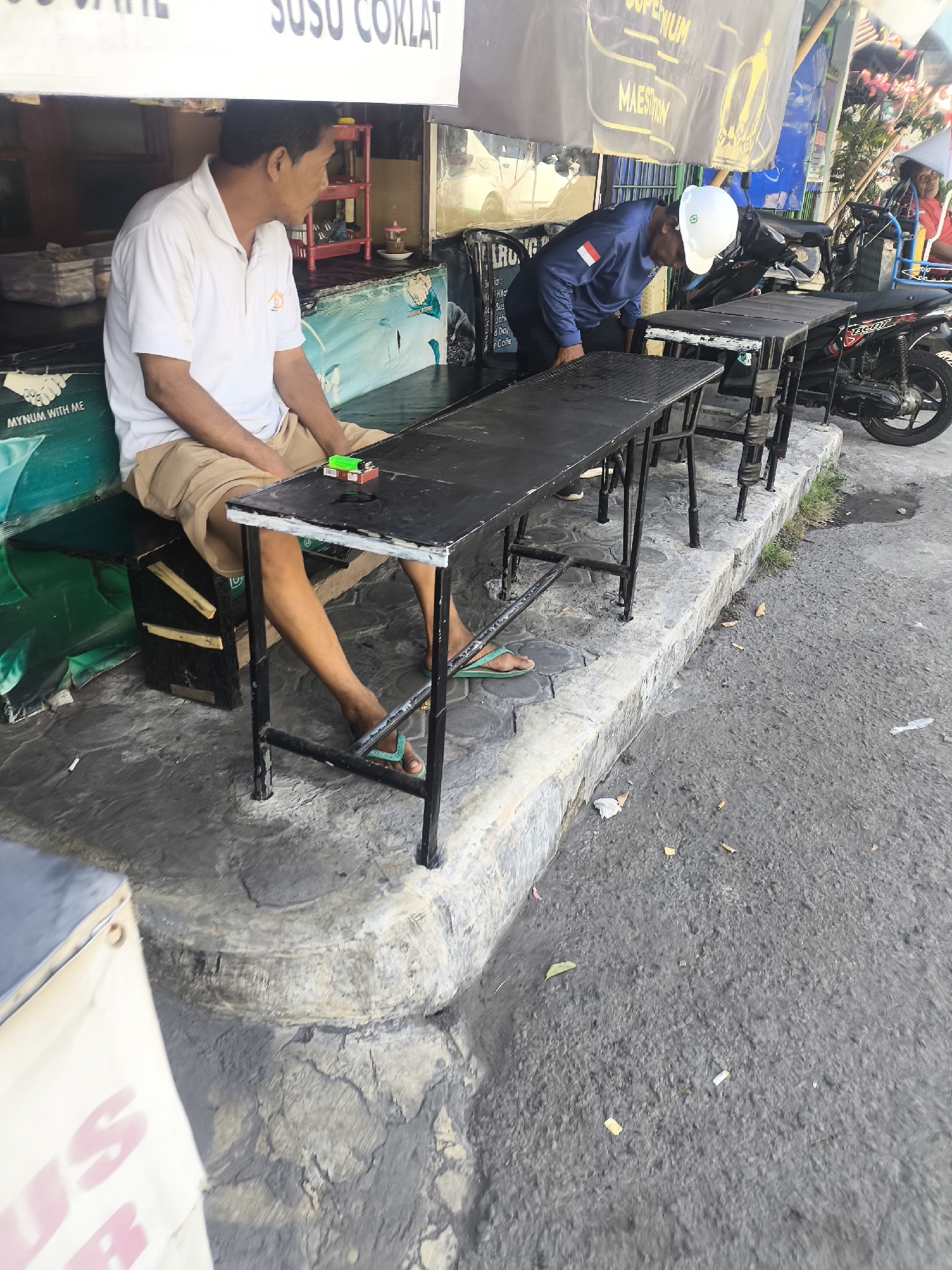 In Lumajang, Indonesia, a man in a white polo shirt and khaki shorts sits on a bench, looking to his right. In front of him, a black table with metal legs is set up outdoors, beside another identical table. A second man, wearing a blue long-sleeved shirt, white hard hat, and dark pants, bends over the second table, possibly arranging items. A black motorcycle is parked behind the tables. The ground is paved with textured concrete and asphalt, littered with cigarette butts and debris. A white banner with blurry red lettering hangs above the scene.