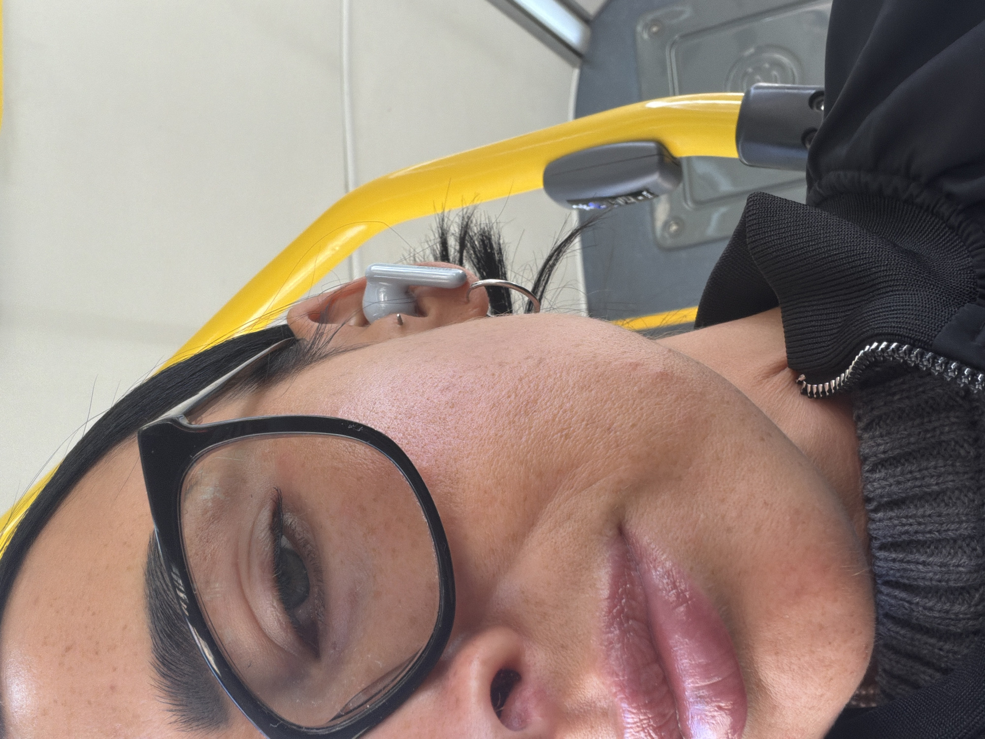 A close-up, tilted photograph depicts a woman wearing black-rimmed glasses and a jacket with a zipper detail. An assistive listening device is visible in her ear. The background includes a yellow handrail and a gray seat, suggesting a public transport setting. Considering the presence of these elements, the photograph likely originates from the city of Saint Petersburg, Russia, due to its extensive public transportation system featuring similar handrails and seat designs.