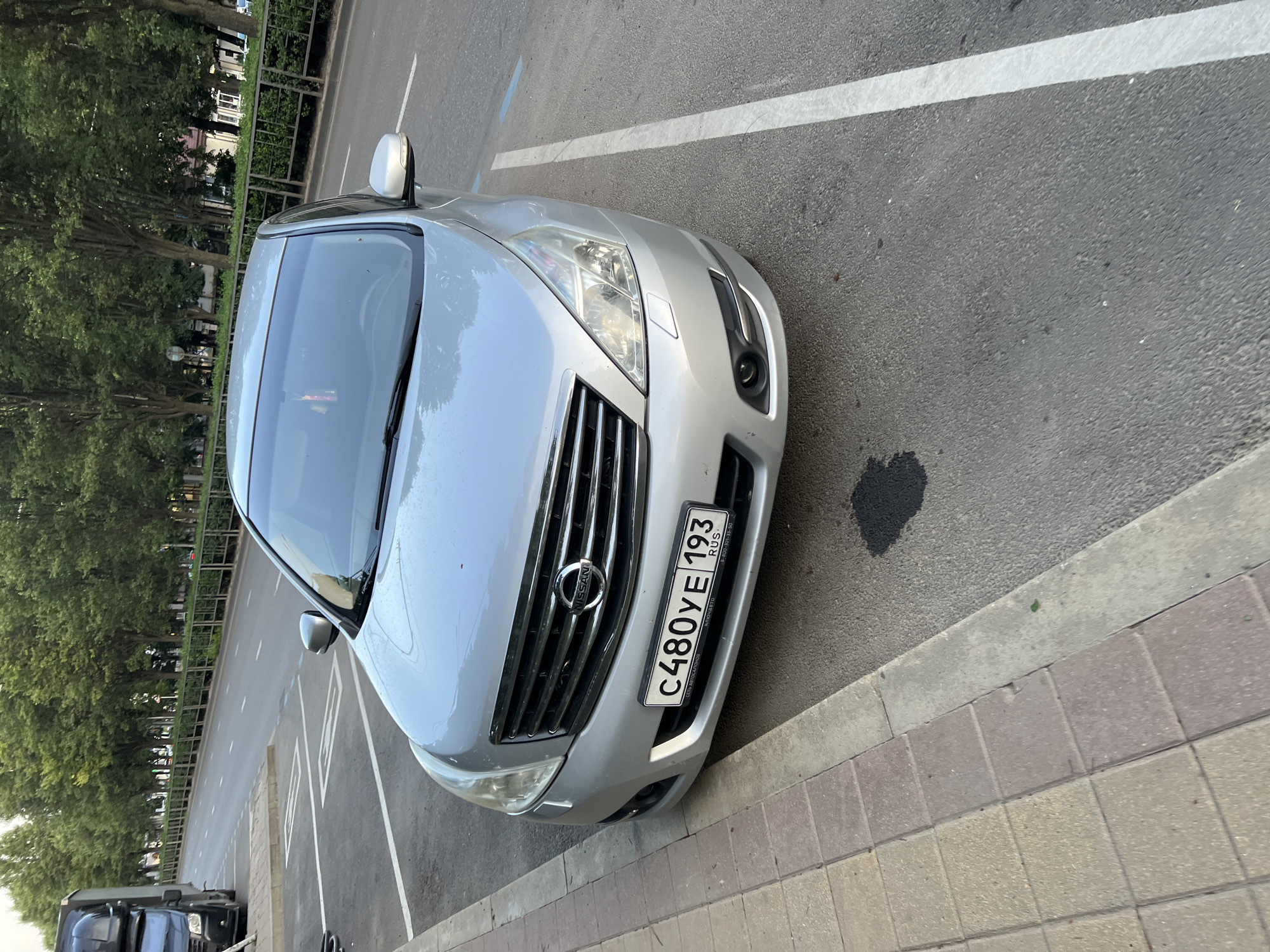 The image shows a silver Nissan Teana parked on a street in Krasnodar, Russia. The car's license plate reads "C480YE 193". The car is parked next to a sidewalk with light brown and grey paving stones. The road is paved with asphalt and has white lane markings. In the background, there are trees, a metal fence, and buildings. A black truck is visible parked on the left side of the image, partially obscuring the street. A dark fluid stain is visible on the asphalt in front of the Nissan. The sky is overcast.