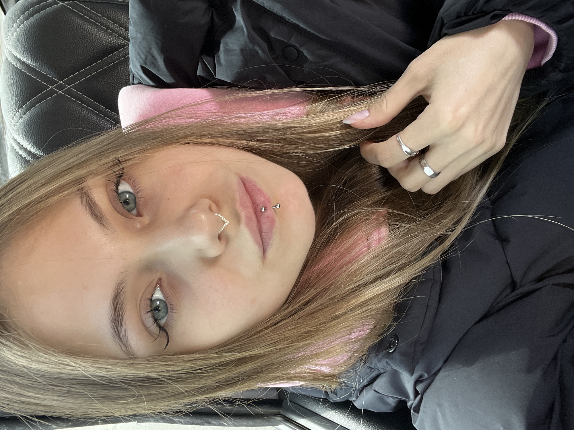 A close-up, rotated portrait depicts a young woman with long, straight light brown hair, fair skin, and light-colored eyes (appearing blue or grey). Her expression is neutral to slightly pensive as she looks directly at the camera. She is positioned with her head angled, possibly lying down or leaning back.
She is wearing a black puffer jacket, with a soft pink lining visible around her neck and at the cuff of her right sleeve (based on an upright orientation). Her left hand is gently placed in her hair, near her head. This hand features manicured nails with a natural, light polish and two simple silver-toned rings on her index and middle fingers.
The woman has distinct facial piercings: a V-shaped, silver-toned septum ring with small embellishments on her nose, and a philtrum piercing (medusa piercing) above her upper lip, consisting of two small silver studs. Her eyebrows are well-defined, and her eyelashes appear long.
The background primarily consists of a quilted black surface with white stitching, suggesting the interior of a vehicle, such as a car seat, or a similarly upholstered piece of furniture. The lighting is soft and even, indicating an indoor setting, and the time of day is not discernible. No visible text is present in the image. The geographical context provided for the image is Фоки, Russia.