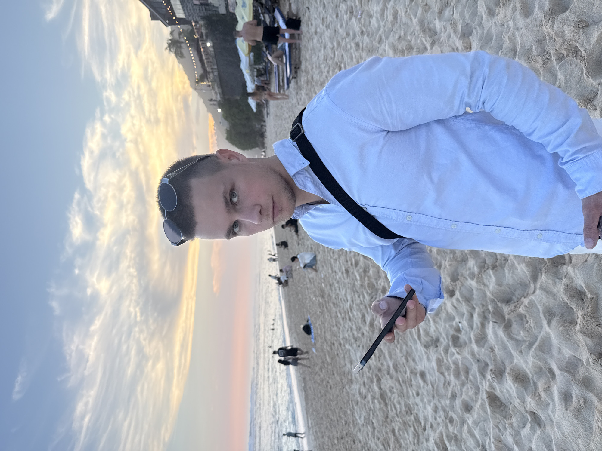 A man is standing on the beach. The man is wearing a white button-down shirt and has sunglasses on his head. He is holding a black phone or similar electronic device in his right hand and a black strap is diagonally across his body, over his left shoulder. The beach is sandy and there are people in the background both on the beach and in the water. There is a building on a hillside in the distance. The sky is cloudy and appears to be around sunset or sunrise. The location appears to be on the beach in Kuta Selatan, Indonesia.