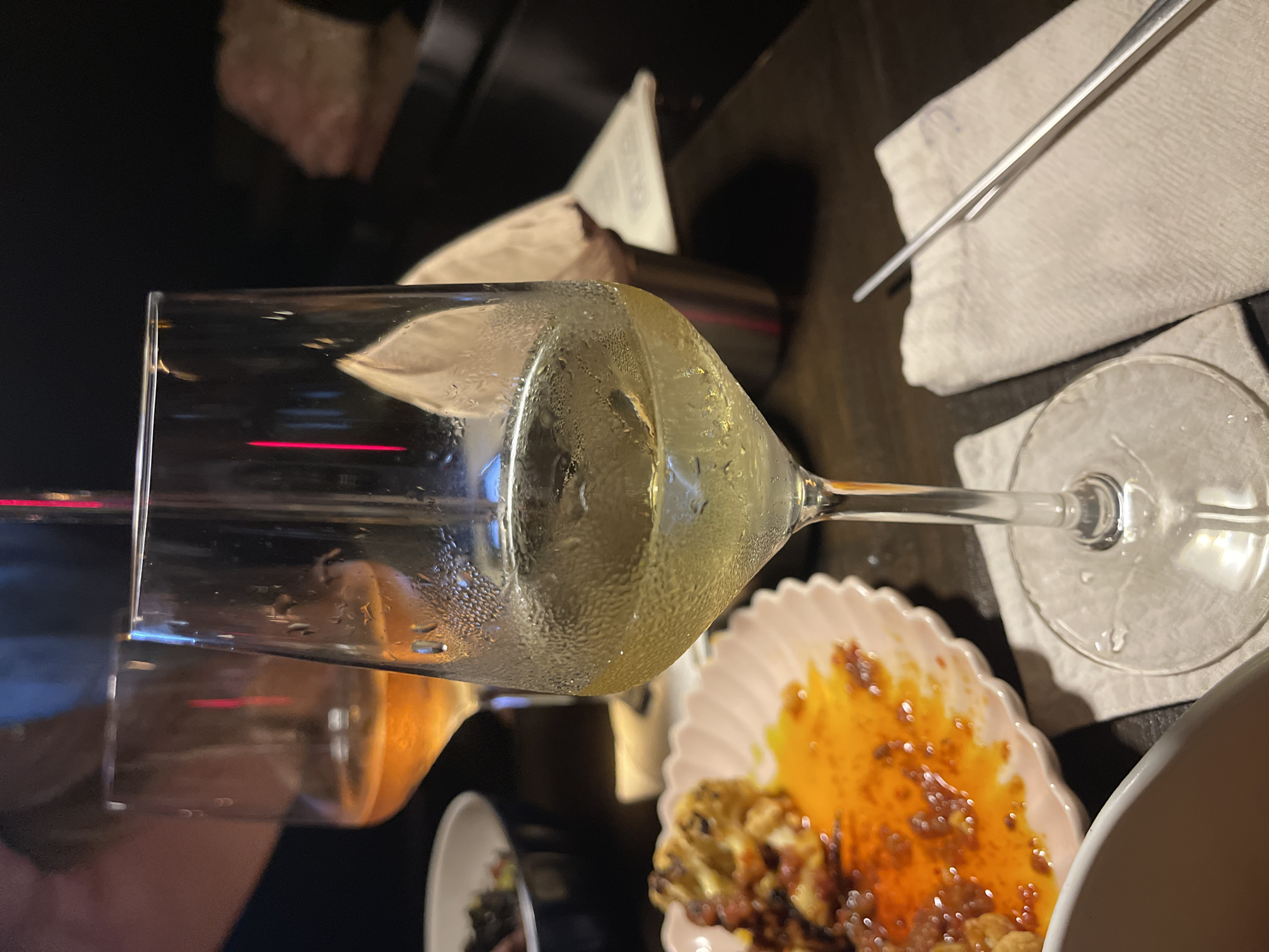 The image shows a close-up, tilted view of a wine glass filled with a pale yellow liquid, possibly white wine. The glass is sweating, with numerous small water droplets clinging to its surface, suggesting it is chilled and the ambient temperature is warm. This detail, along with the presence of food on a decorative plate in the foreground, implies a meal or dining experience is taking place.
In the background, out of focus, there are hints of a dark, possibly wooden table. A white napkin or cloth is visible, with a pair of silver chopsticks resting on it. The lighting appears to be artificial, creating reflections and highlights on the glass and plate. The overall impression is one of a relaxed, possibly celebratory or social occasion.
The setting seems to be an indoor dining establishment, given the table setting and the overall ambiance. The colors are muted, with the dominant tones being the pale yellow of the wine, the orange hues of the sauce on the plate, and the dark background. No people are directly visible, but the presence of cutlery and drinks suggests their recent or imminent presence. The image does not contain any visible text.
Considering the provided location context of Mengwi, Indonesia, the presence of condensation on the glass is consistent with the tropical climate. The meal appears to involve some sort of saucy dish, and the chopsticks suggest Asian cuisine may be part of the dining experience. The overall mood evoked is one of enjoyment and indulgence.