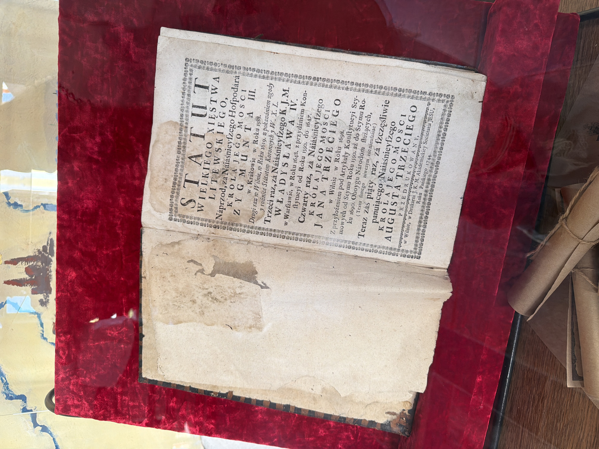The image shows an open, aged book with yellowed pages displayed within a red velvet-lined casing, likely for preservation or exhibition. The book's title page is visible, featuring elaborate decorative borders and Polish text. The text is in an old script, suggesting it is a historical document. The surrounding environment appears to be indoors, with a wooden surface beneath the display. There are no people visible in the image. The context of "Літвенскі сельскі Савет, Belarus" is not directly discernible from the visual information presented in the image itself.
