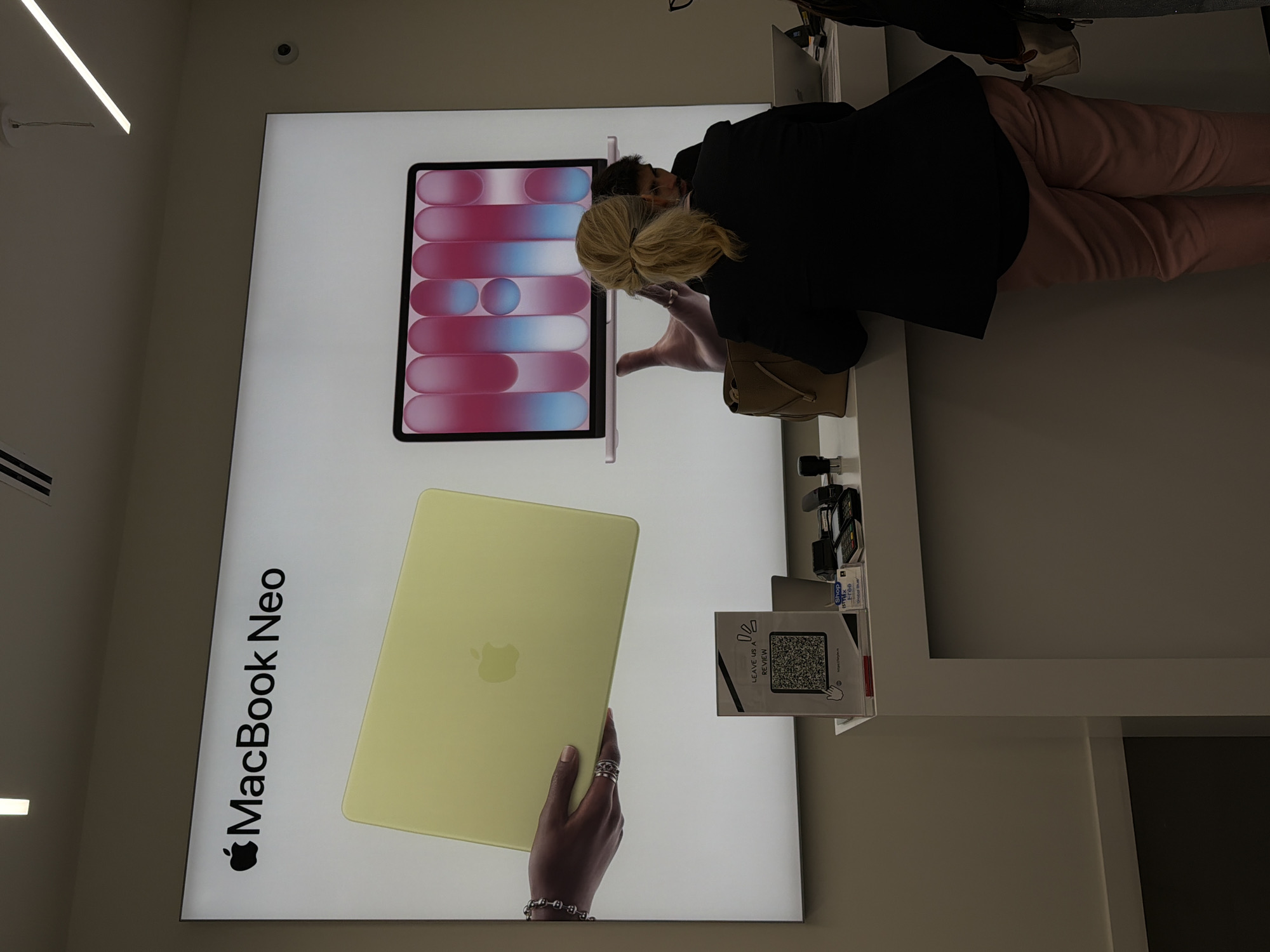 In a retail environment, likely an Apple Store or a similar electronics retailer, a large backlit display features the text "MacBook Neo" with an Apple logo. Below the display, a white counter is visible. On the counter, there are several items, including payment terminals, a paper stand with a QR code and the text "LEAVE US A REVIEW," and a laptop. Two individuals are positioned behind the counter, interacting with the merchandise or engaging with customers. One individual, a woman with blonde hair pulled back in a ponytail, is wearing a black blazer and pink pants. Another individual, a man with dark hair, is partially visible. In the foreground, the lower half of the counter and the floor are visible, with a white baseboard running along the wall. The overall impression is that of a modern retail setting.