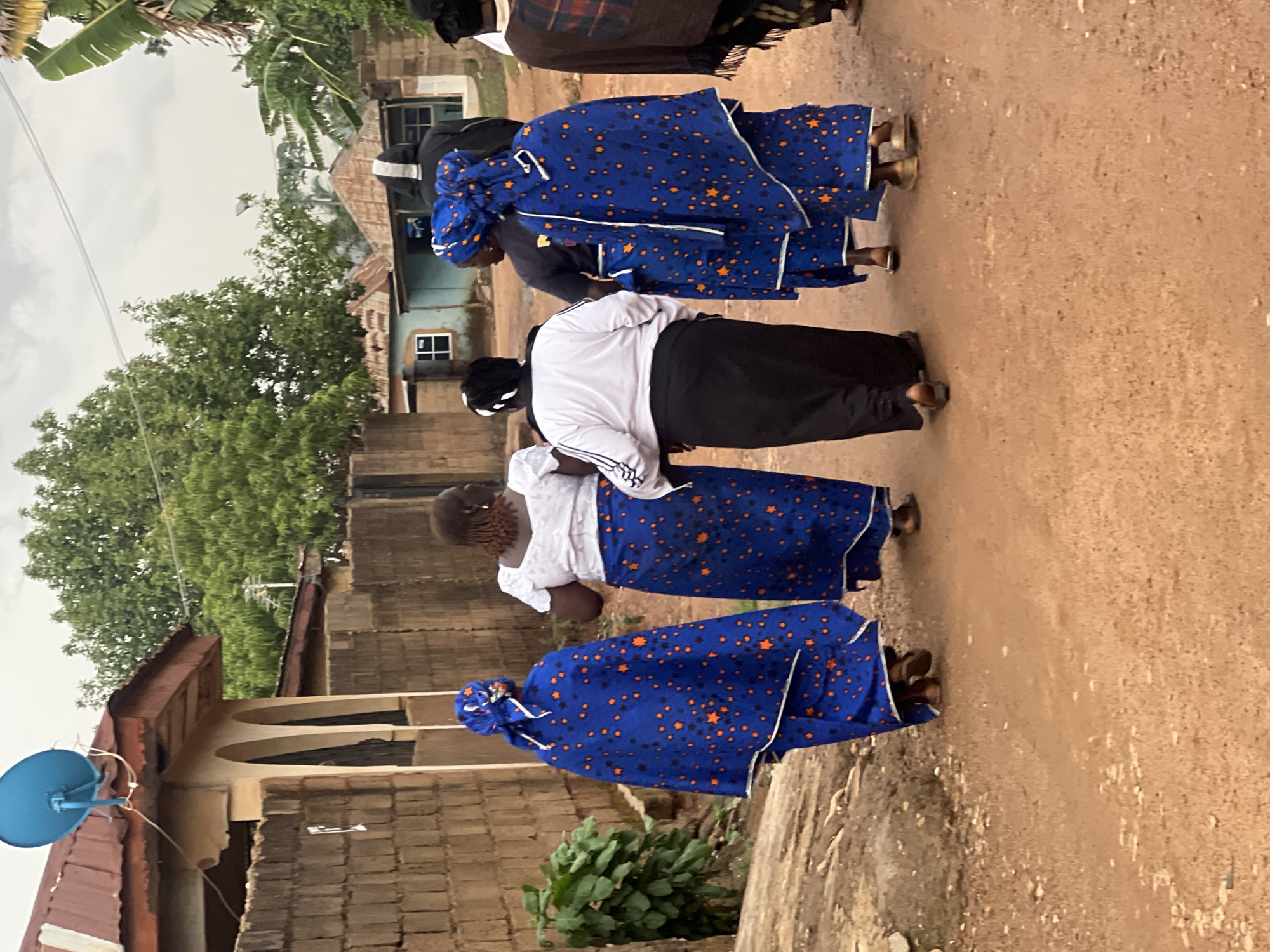 A row of women walk on a dirt road in Gura Top, Nigeria. The women are dressed in blue fabric decorated with orange stars. One woman wears a white top and black skirt, with a white head covering. The surrounding area includes residential buildings constructed from brick and concrete. A blue satellite dish is affixed to the roof of one of the buildings. Green vegetation and trees are also present. The sky is overcast.