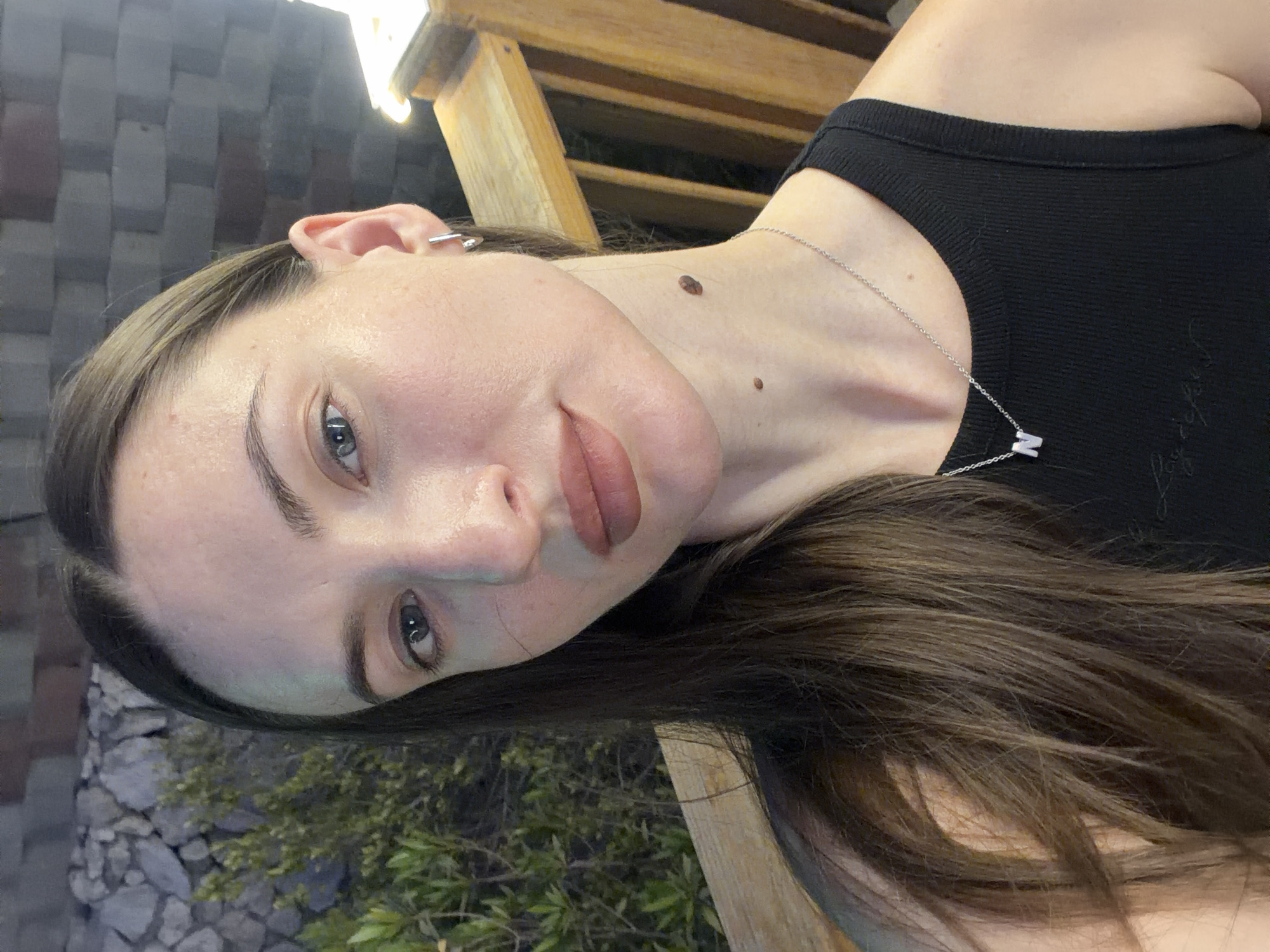 A woman with long, dark brown hair is posing for a selfie outdoors at night. She is wearing a black tank top and a silver necklace with the letter "N" pendant. She has a mole on her neck. The background appears to be a wooden railing and some foliage, with a stone wall further behind. The lighting suggests an artificial light source. The location is not specified in the image or its context.