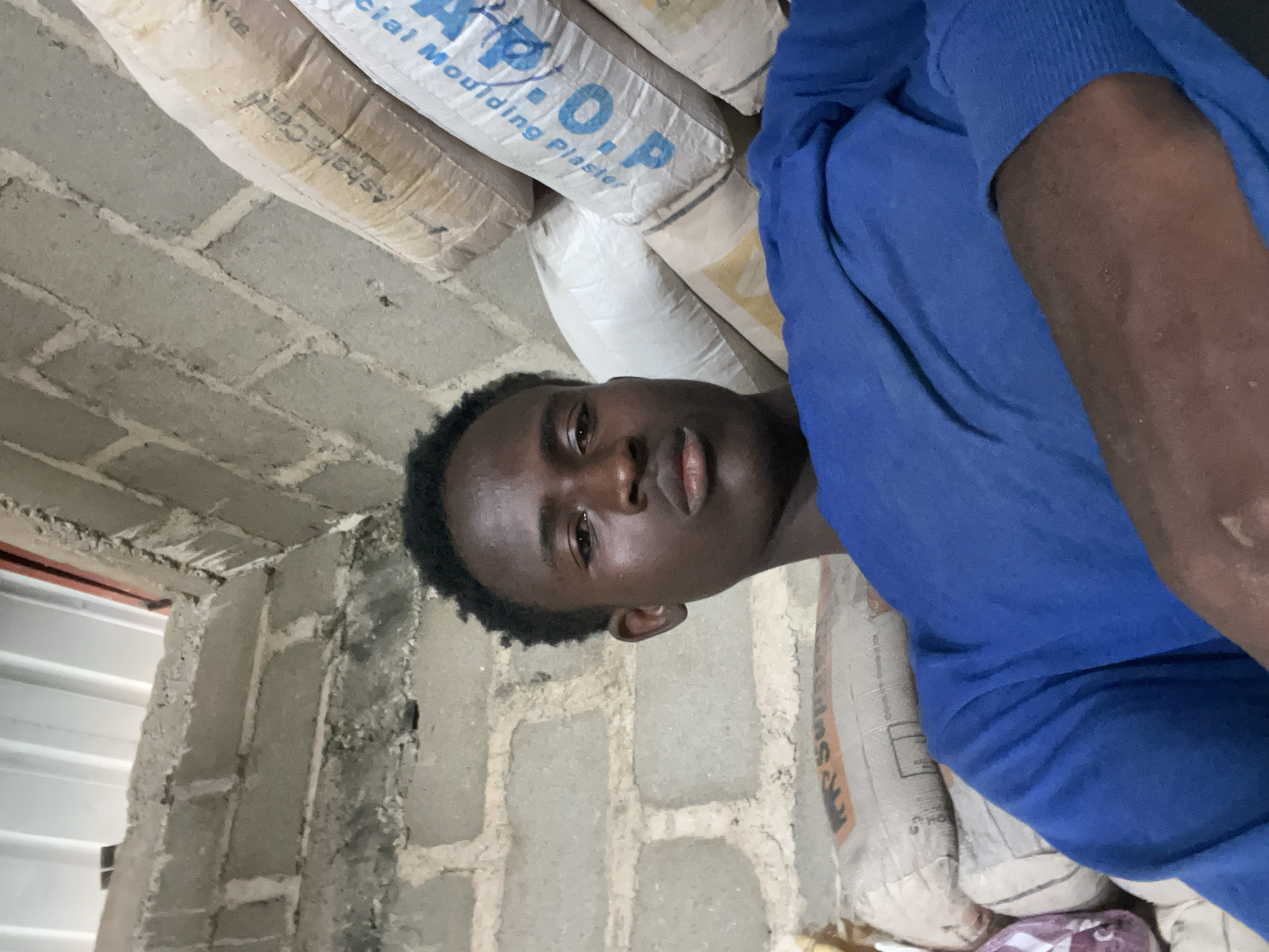 A young Black male is pictured in a close-up selfie, seated indoors with a pile of bagged materials, possibly cement or plaster, stacked behind him. He is wearing a blue long-sleeved shirt. The background consists of unplastered brick walls and what appears to be a corrugated metal ceiling or roof structure. The lighting suggests an indoor setting, possibly a storage area or construction site. The image is focused on the young man's face and upper body, with the bags and walls providing a textured backdrop. The overall impression is a candid shot taken in a utilitarian environment.