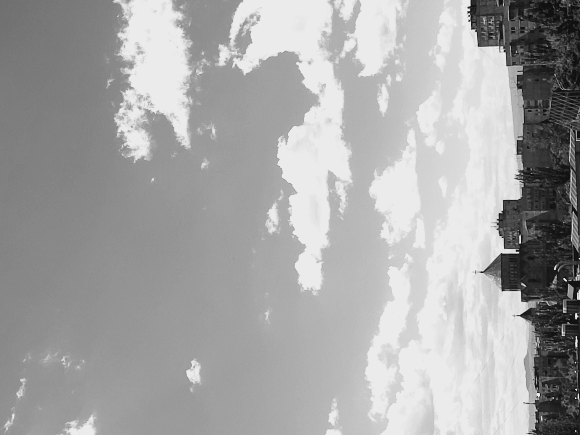 The image, rotated 90 degrees counter-clockwise, shows a partial view of the city of Yerevan, Armenia, as seen against a cloudy sky. Buildings line the horizon, varying in height from low-rise structures to taller apartment blocks. Church spires are visible, interspersed among the buildings. Trees and other vegetation appear scattered throughout the urban landscape. The sky dominates the composition, filled with various cloud formations. The entire image is in black and white, adding a vintage or documentary feel.