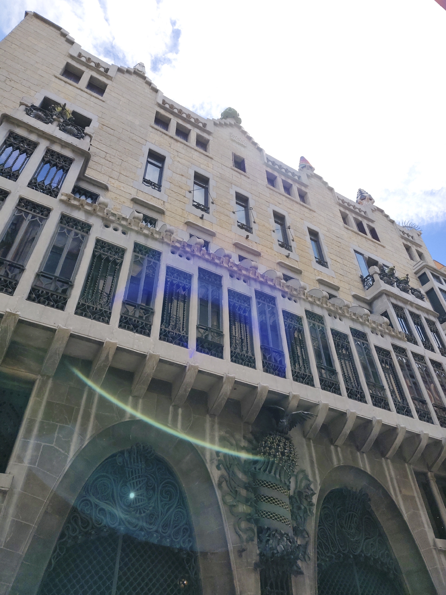 The image depicts the exterior of Casa Batlló, a renowned building located in Barcelona, Spain. The building is characterized by its Modernist architecture. The facade is predominantly light stone. It features a series of arched windows on the ground level, covered with ornate, wrought-iron designs. The upper levels have larger windows with similar decorative ironwork. Some balconies are visible, adorned with plant life. At the top of the building, there are decorative elements, including a green tiled spire. The sky is partly cloudy. The viewpoint is from below, looking upward at a steep angle.