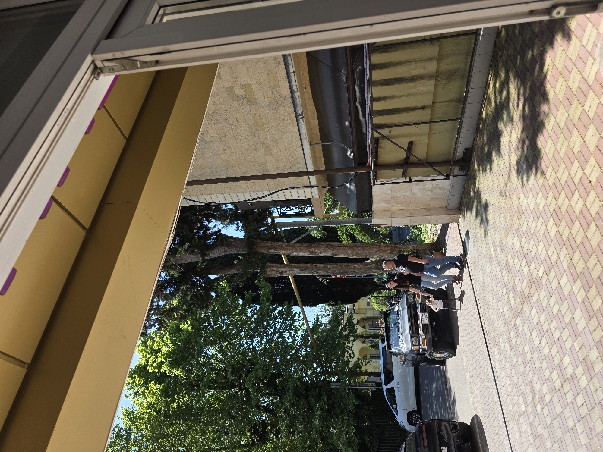 The image, rotated 90 degrees clockwise for proper viewing, depicts an outdoor scene on a sunny day, likely in Sochi, Russia. In the immediate foreground, part of an open window frame, featuring grey aluminum or uPVC profiles and hinges, is visible on the left.
The main focus of the scene is a paved sidewalk or street area with distinctive red and tan interlocking pavers. Standing on this pavement, near a parked grey SUV, are two adults and a young child. The adult male wears a dark t-shirt and jeans, and the adult female wears a dark t-shirt and lighter-colored pants. The child, in light clothing, stands in front of the SUV. The SUV displays a Russian license plate "T 437 EH 193". Behind it, a white van is parked, and a dark vehicle is partially visible in the bottom right corner with a license plate starting "P 242".
The background is rich with vegetation, including tall, dark green cypresses and other lush green trees, some appearing to be palms, indicative of a warm climate. Several buildings line the street; one on the left has a yellow or tan facade, while others further back feature stone or brick-like exteriors and large windows. Overhanging structures, possibly awnings or balconies, are also present. The clear blue sky and strong shadows cast by trees and structures across the pavement suggest bright sunlight during the day. The scene portrays a casual moment in an urban or suburban setting.