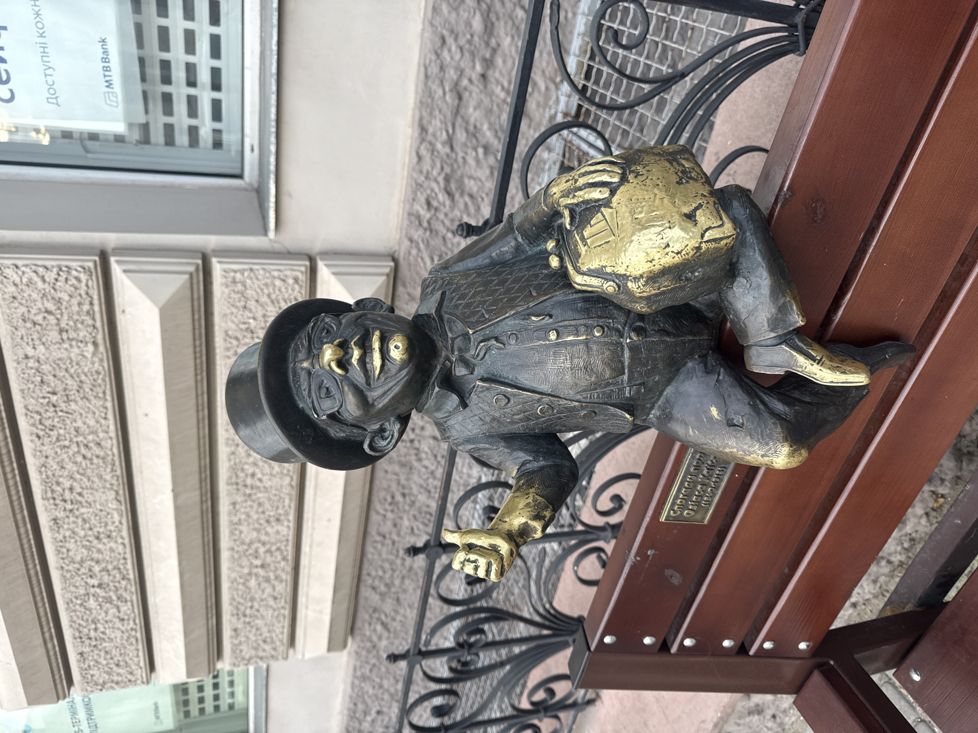 A bronze statue of a dwarf wearing a black top hat and glasses sits on a wooden bench. The dwarf is dressed in a dark suit with a vest and is holding a golden briefcase in its left arm, while its right arm is raised with a thumbs-up gesture. A small plaque is attached to the bench. In the background, a building with a stone facade and windows is visible, one of which has the logo "MTB Bank". The scene is located in the city of Odesa, Ukraine.