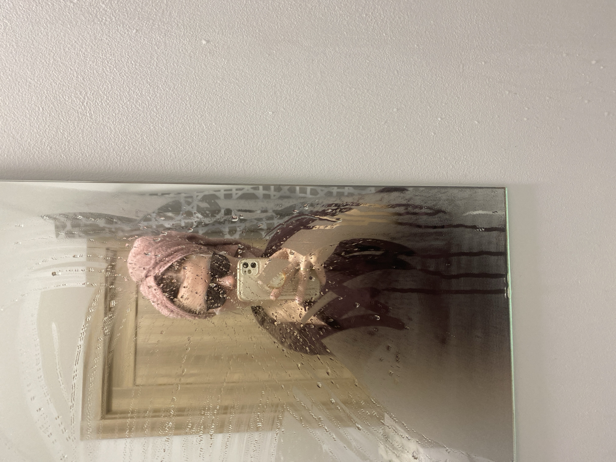 A person with a pink towel on their head takes a mirror selfie in a bathroom. The mirror is steamy with water droplets running down the surface, obscuring the reflection. The wall adjacent to the mirror is painted white. The city of Yekaterinburg, Russia, is not evident in this image.