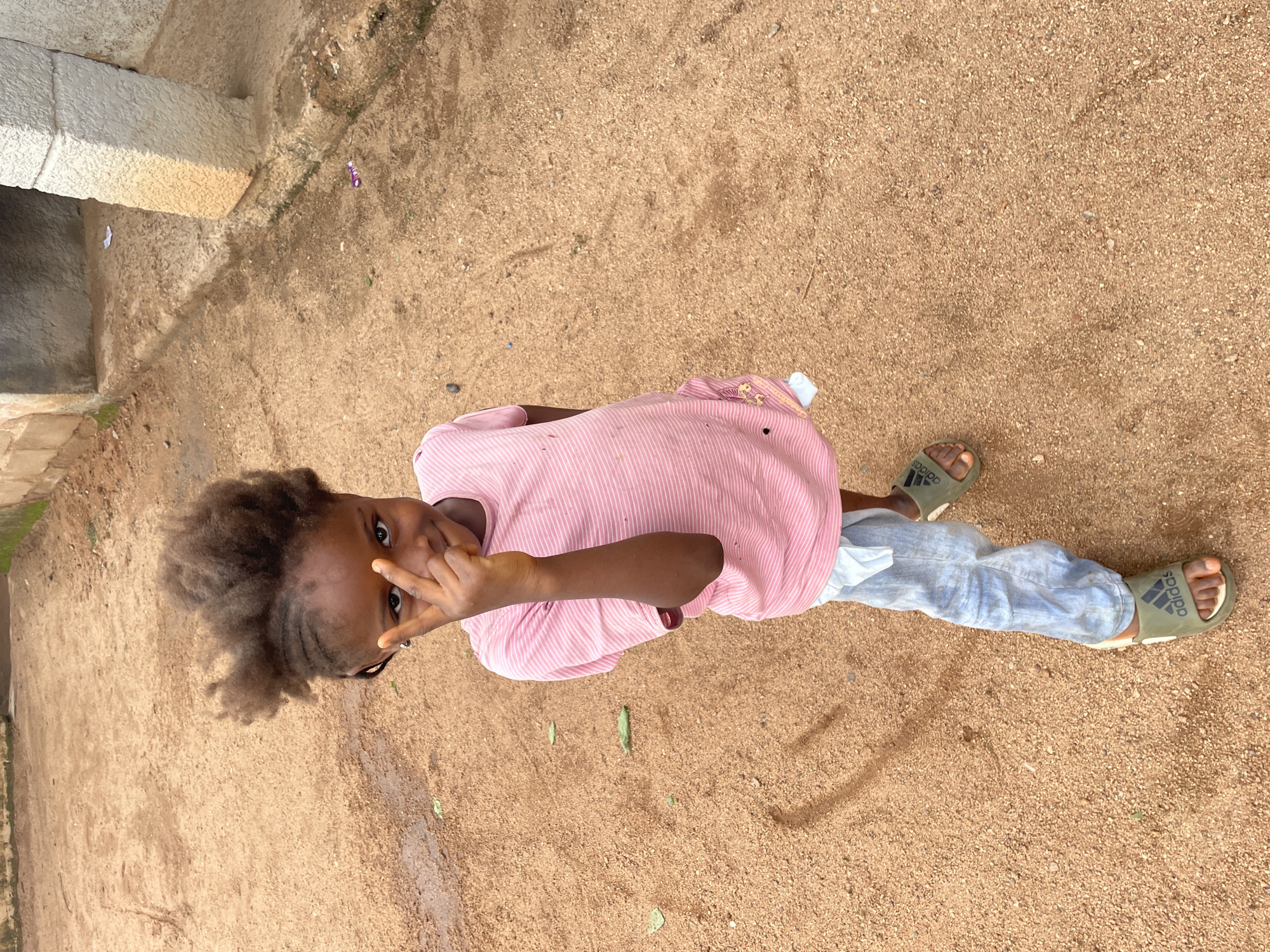 A young girl with dark skin and curly hair, styled in thin braids, stands on a sandy surface in Barakin Lerawi, Nigeria. She is wearing a pink, horizontally striped t-shirt and light blue jeans, with white fabric tucked into the waistband of her jeans. On her feet are green Adidas slides. Her right hand is raised, with her index and middle fingers extended in a peace sign, held near her face. She is looking directly at the camera with a slight smile. The background consists of a sandy, uneven ground with some scattered leaves and debris. To the right, a portion of a weathered concrete structure with a large stone pillar is visible. The lighting suggests it is daytime.