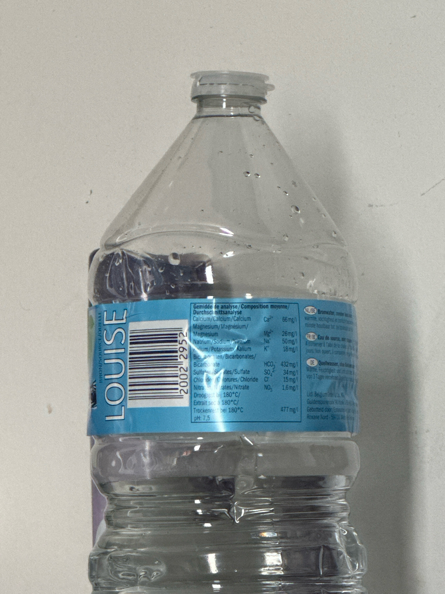 A clear plastic bottle of Louise water is shown upright, against a light-colored, textured background. The bottle has a blue label with text and a barcode printed on it. The label also displays a nutritional analysis in multiple languages, listing the mineral content of the water. The water level is visible, filling approximately the lower third of the bottle. The cap of the bottle is off. The location in Zele, Belgium is mentioned on the label as "Roxane Nord - 59110 Wez", although this is a French address, not Zele, Belgium.