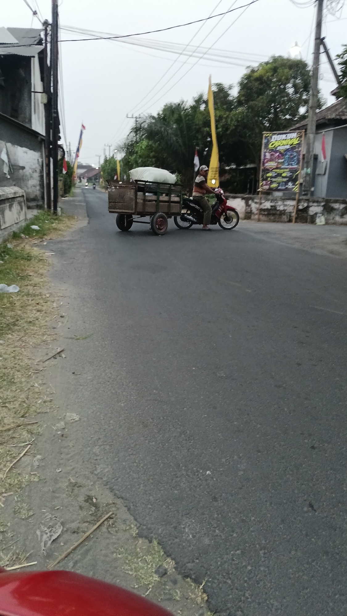 A person is riding a red motorcycle with a trailer attached on a paved road in Cendono, Indonesia. The trailer is filled with goods covered by a large white sheet. The person is wearing a striped shirt and dark pants, and a cap. In the background, there are trees, buildings, and utility poles with wires. A yellow flag and two Indonesian flags are visible. A billboard advertising "DUKUN CARNAVAL" is also present. The sky is overcast. The road appears to be a street in a residential or semi-rural area.