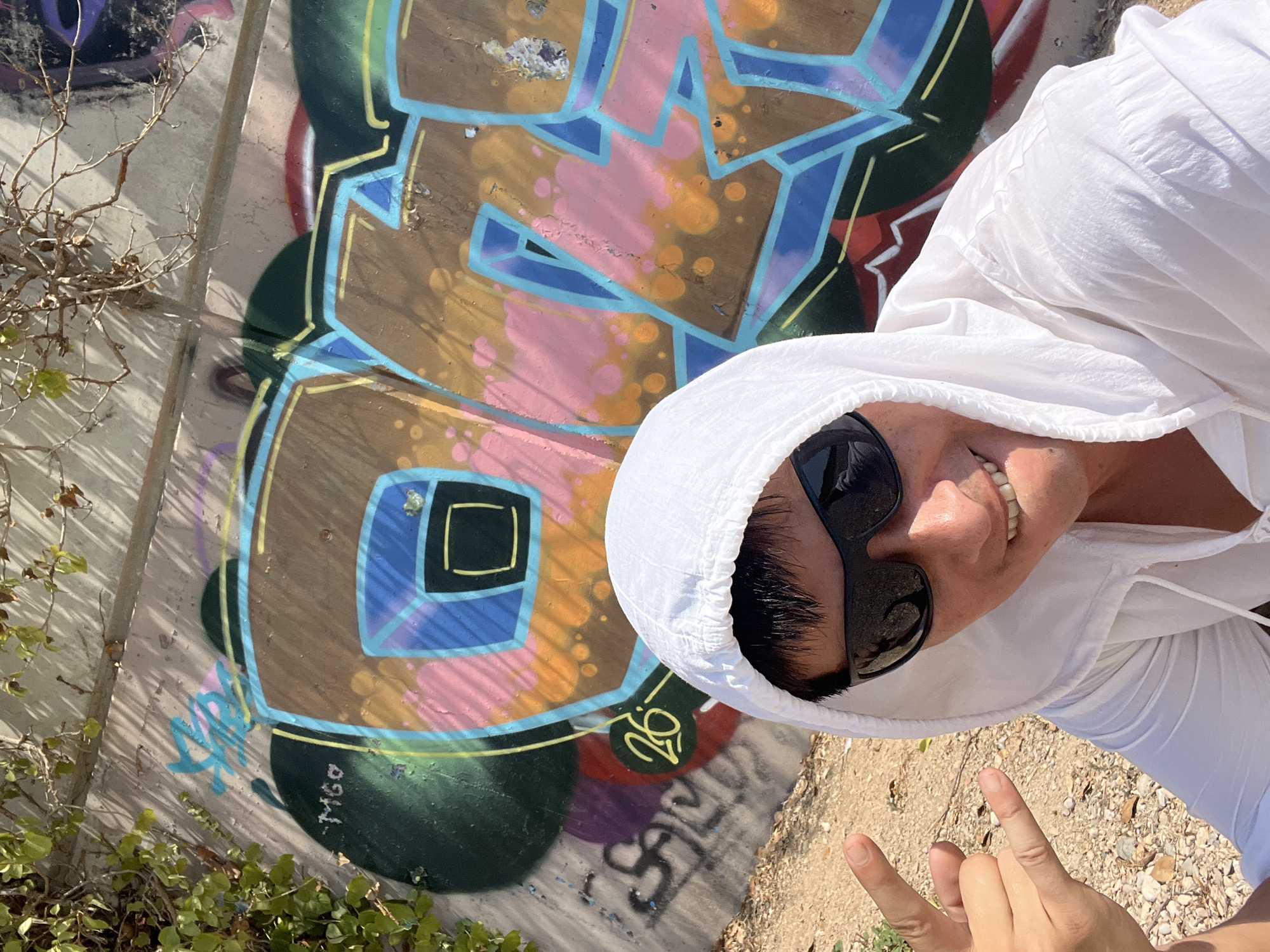 A man wearing sunglasses and a white hooded jacket is taking a selfie in front of a graffiti-covered concrete wall. The graffiti features bold lettering in various colors, including brown, blue, pink, and orange. To the left of the man, his hand is raised, making a rock sign with his index and pinky fingers extended. The ground is covered with gravel and some sparse vegetation. The background suggests an outdoor urban or semi-urban setting. The graffiti includes tags like "M60" and "26". The overall scene appears to be outdoors, possibly in an area with public art or urban decay, consistent with locations in Tarragona, Spain, known for its street art.