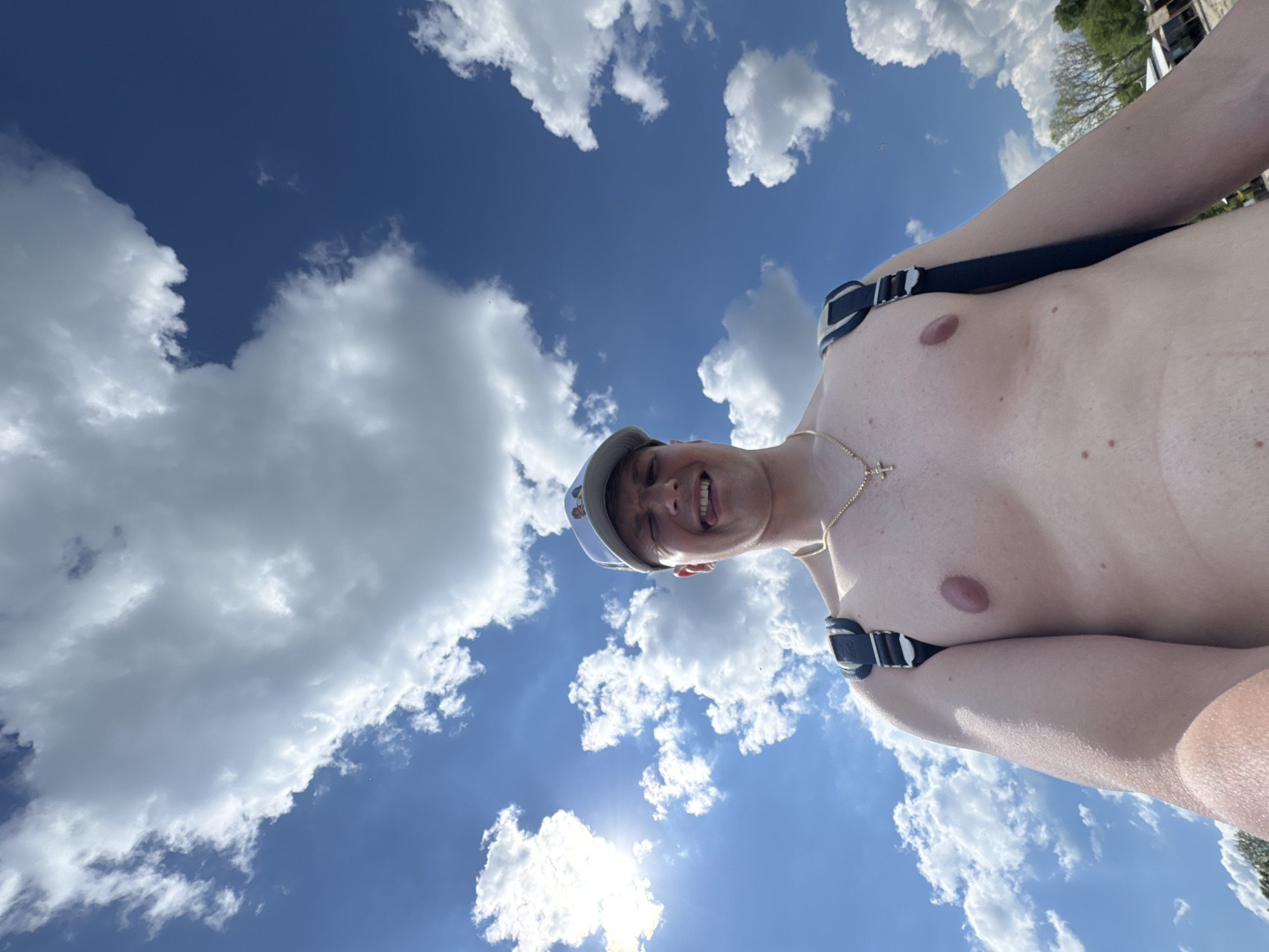 A low-angle, upward-tilted shot captures a shirtless person against a backdrop of a blue sky with fluffy white clouds and a bright sun. The individual, possibly male, is wearing a baseball cap and a thin gold chain with a cross pendant. They are wearing what appears to be a strap or harness over their shoulders. The location might be in the city of Burgas, Bulgaria, but there is no clear indication of landmarks or characteristics specific to that location in the image to confirm it.