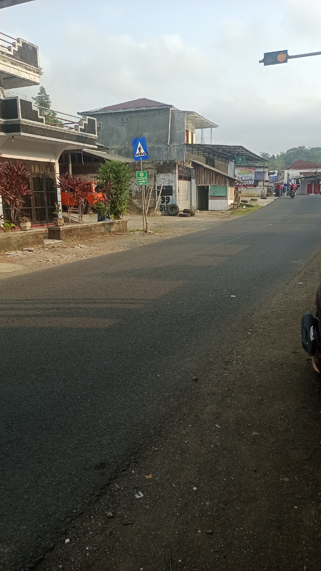 A street scene in Karyamandala, Indonesia, shows a paved road curving towards the right. On the left side of the road, there is a building with red plants in front of it and an orange vehicle parked behind a fence. Further down the road, there are several buildings, including one with a grey facade and a red roof, and another with an open front displaying various items. A pedestrian crossing sign is visible on the left side of the road, and a traffic light with an amber signal is positioned in the upper right corner of the image. Several people on motorcycles are seen on the road in the distance. The sky is clear with some clouds, and the overall atmosphere suggests it is daytime.
