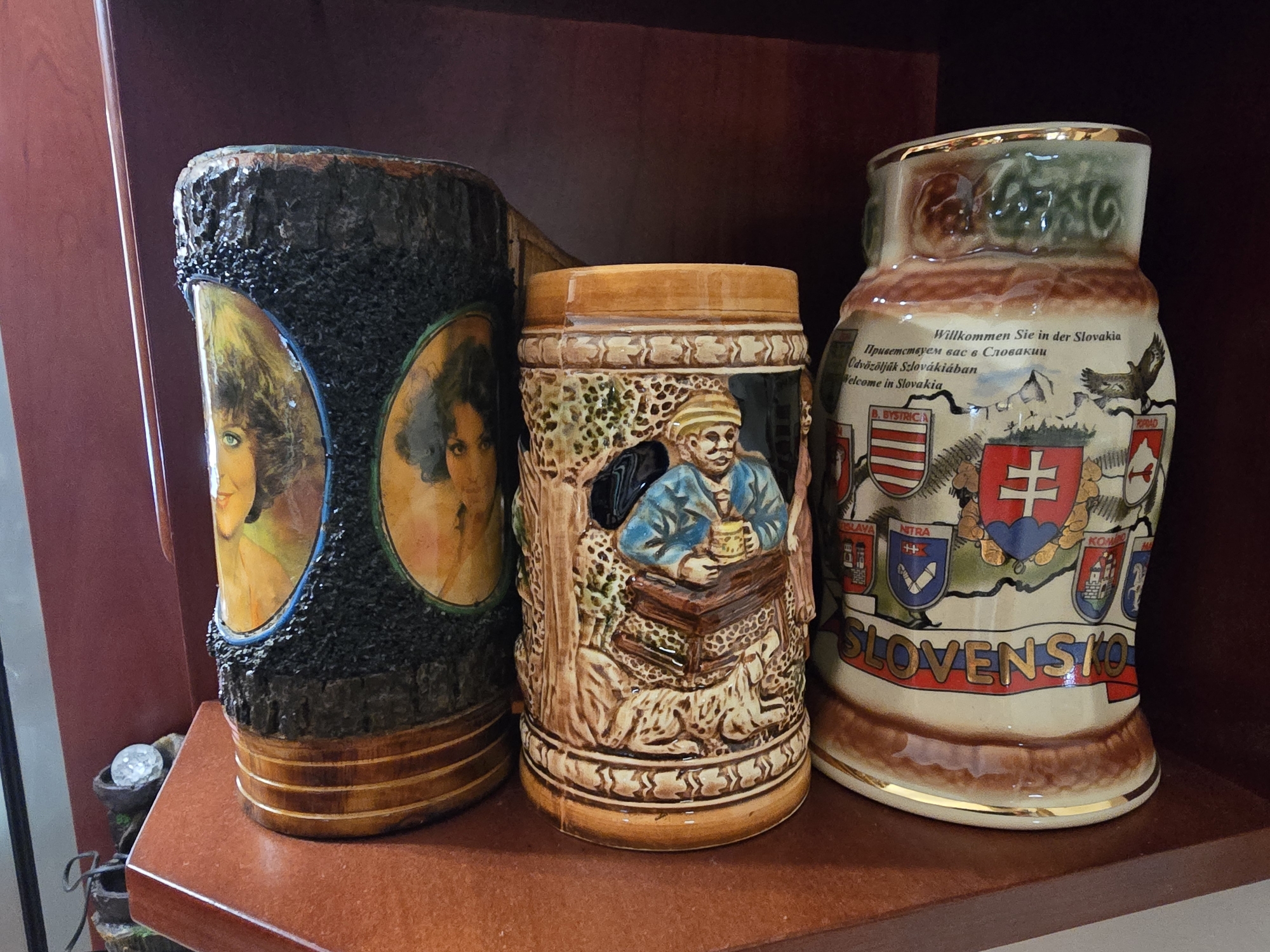 The image displays three decorative ceramic mugs or steins arranged on a reddish-brown wooden shelf. The shelf is positioned within a dark wooden cabinet.
From left to right:
1. The first object is a cylindrical mug with a dark, textured exterior resembling tree bark. It features two oval insets, each depicting a woman's portrait with fair skin and dark hair. The woman on the left has green eyes, and the woman on the right has dark eyes. The mug has a wooden base with horizontal bands and a gold rim.
2. The middle object is a beer stein with a detailed bas-relief scene. It depicts a man wearing a blue shirt and a striped head covering, sitting and holding a small mug. He is positioned behind what appears to be a wooden surface or table. The stein is primarily brown and cream-colored, with some green accents. It has a light brown rim.
3. The third object, on the right, is a tall, tapered stein with a cream-colored base decorated with various images and text. The text includes "Willkommen Sie in der Slovakia" in German, "Приветствуем вас в Словакии" in Russian, and "Welcome in Slovakia" in English. The stein features coat-of-arms, a map outline of Slovakia, and images of buildings and a bird in flight. The word "SLOVENSKO" is prominently displayed in gold lettering across the base. It has a decorative gold rim.
To the left of the objects, a portion of a small decorative item is visible, appearing to be a small tiered fountain with a clear glass sphere at the top. The background of the image is a dark, reddish-brown wooden surface, likely part of the cabinet. The lighting appears to be coming from the front, casting some shadows. There is no indication of the city of Ilion, Greece, in this image.